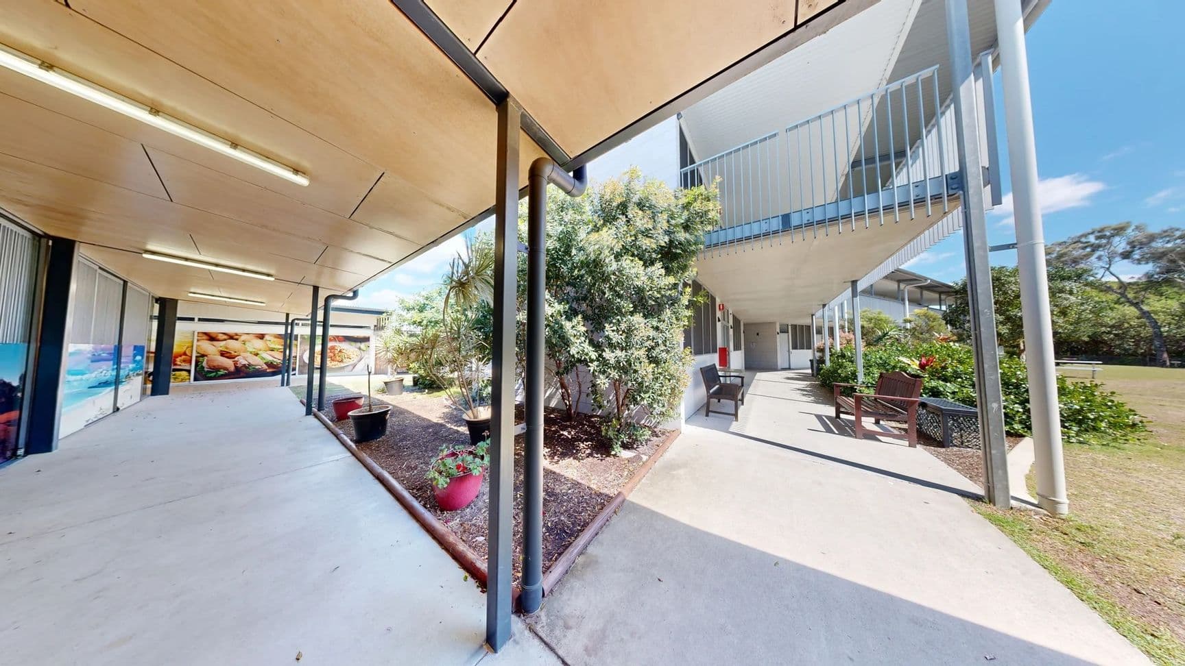 On the left, there's a two-storey building with a balcony containing several doors leading to rooms, alongside a wooden bench. The floor is grass, which is largely uniform and green. On the right, another section of the building is visible, with some greenery and trees nearby. There are picnic tables set on the grass, suggesting an outdoor area for gatherings. The ceiling is not visible, but the structure is modern with a flat roof.
