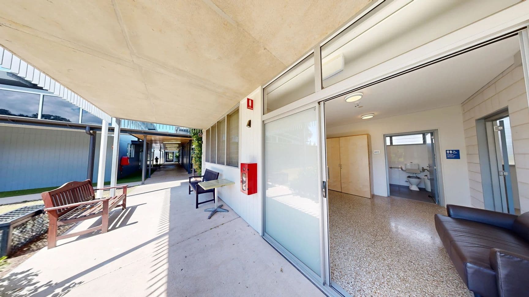 On the left, there is a covered walkway with a bench made of dark wood and a chair beside a small table. The floor is a smooth concrete surface, leading towards an open door revealing a room with light-coloured walls and a polished, light-coloured floor. To the right, there is a doorway leading into a bathroom space, showing a glimpse of fixtures. The ceiling is flat and has simple lighting fixtures arranged evenly.