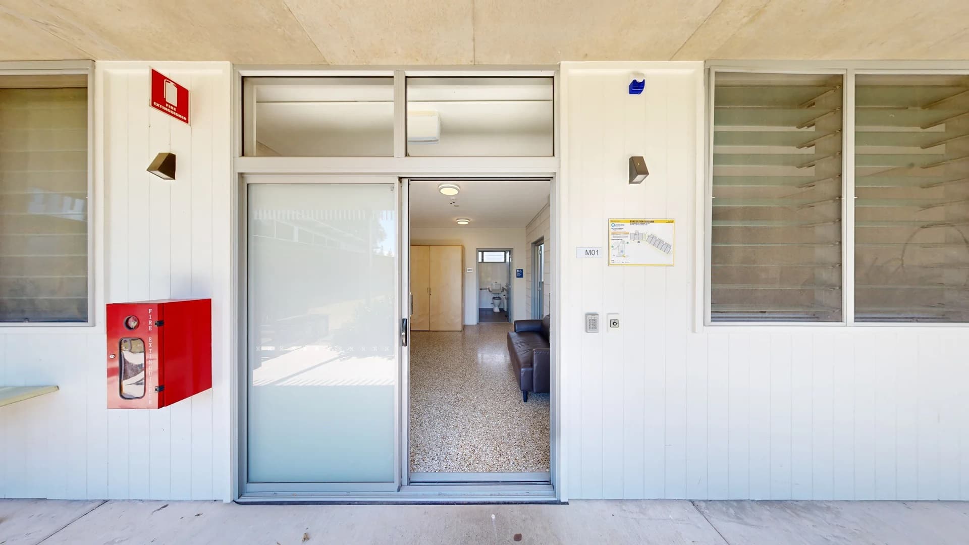 To the left, there is a bright red fire extinguisher box mounted on the wall, and beside it, a small, flat ledge. The entrance features a glass door leading into a well-lit interior with a light-coloured tiled floor. On the right, a series of large windows allow natural light inside, while a wall-mounted light fixture is positioned near the entrance. Inside, a sofa is visible in the foreground, facing an open doorway that leads to a bathroom in the background.