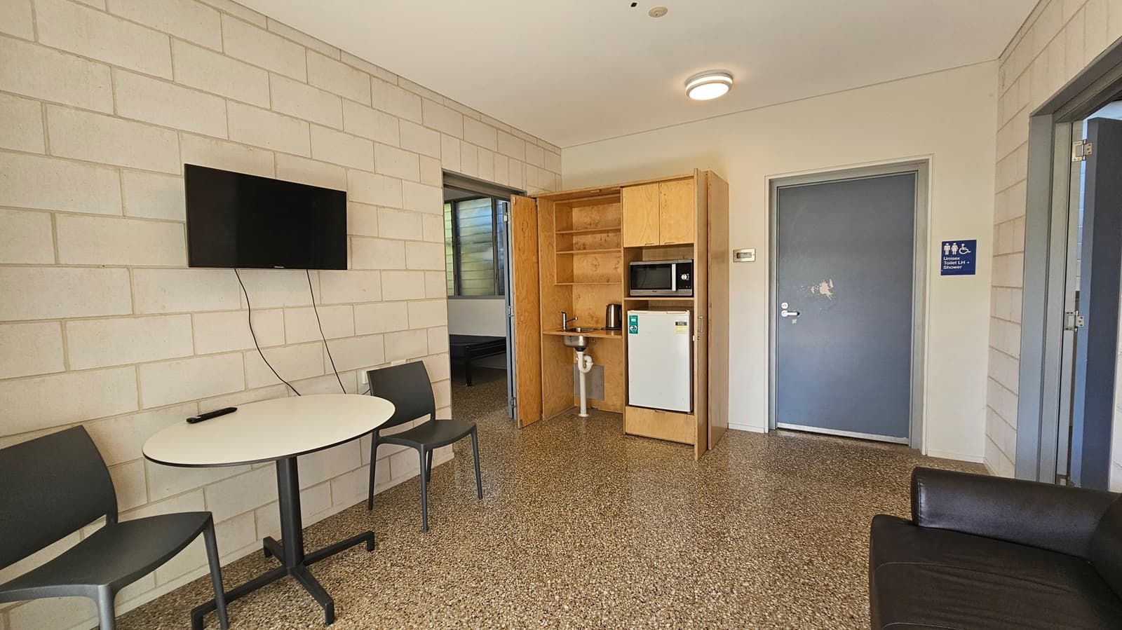 A room with light-coloured brick walls and a speckled floor. On the left, a wall-mounted TV is above a small round table with two chairs. In the centre, a wooden kitchenette features a sink, microwave, and mini-fridge. To the right, a blue door with a sign indicating a unisex accessible toilet. A black sofa is partially visible in the foreground. The ceiling is plain with a circular light fixture.