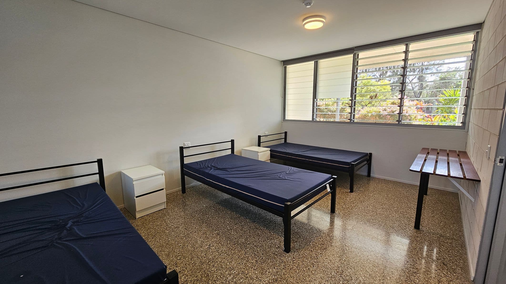 A room with three single metal-framed beds, each with a dark mattress. Two small white bedside tables are positioned between the beds. A large window with horizontal slats is on the right wall, allowing natural light to enter. Below the window is a simple wooden desk attached to the wall. The floor is speckled terrazzo, and the ceiling is plain with a central light fixture.