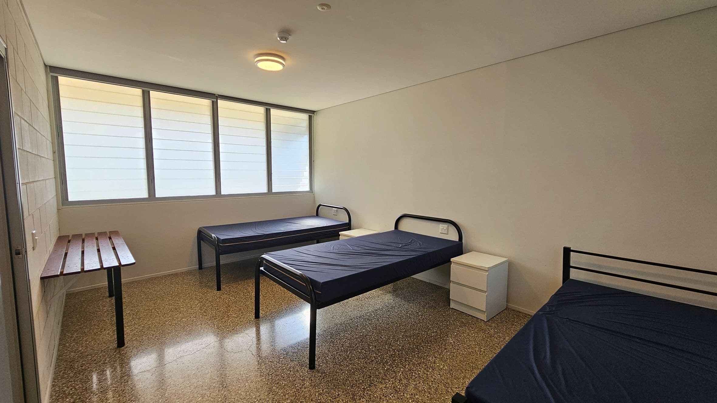 On the left, a wooden shelf rests against the wall, supported by two legs, providing space for personal items. There is a single bed with a dark blue mattress next to it. In the centre, another bed with a similar dark blue mattress occupies the middle of the room, framed by simple metal bed frames. On the right, a small white bedside table with drawers is positioned next to a third bed. The floor is a polished concrete with a smooth finish, and the ceiling is flat with a central light fixture.