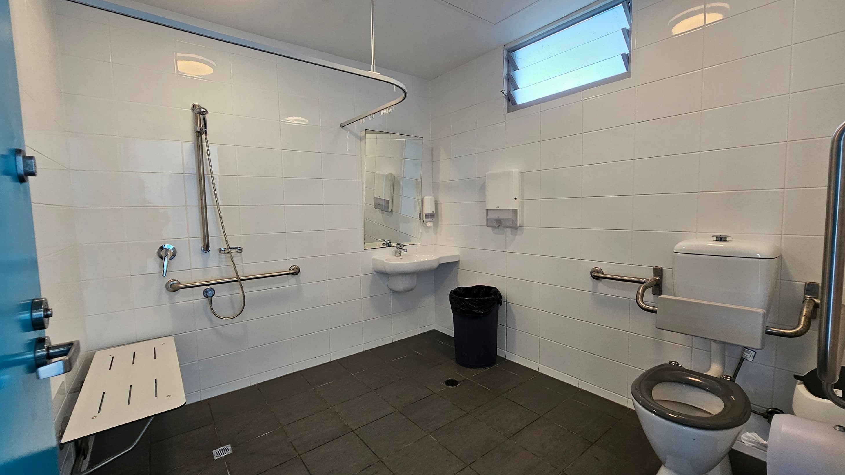 On the left, there is a blue door leading into the restroom, next to a vertical grab bar and a white folding shower seat mounted on the wall. To the right, a toilet is positioned against the wall, equipped with grab bars for assistance. In the centre, a small sink is mounted below a mirror, with a soap dispenser and paper towel holder nearby. The floor is covered in dark tiles, and the white walls have a glossy finish, reflecting light from overhead fixtures and a small window near the top.