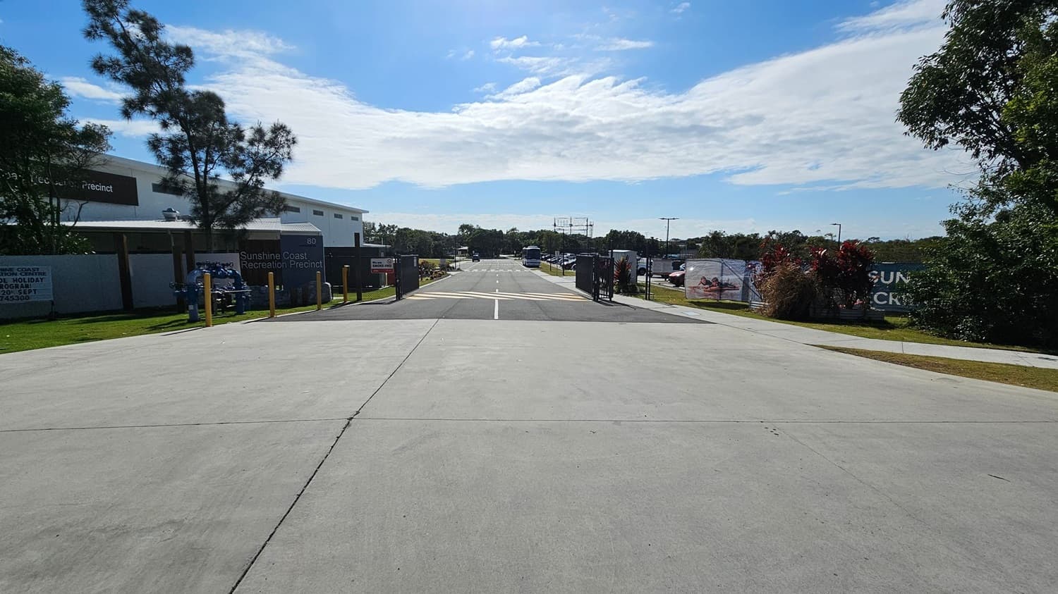 A smooth, concrete pathway stretches down the centre, flanked by greenery on the left and various structures on the right. To the left, there is a low building partially obscured by trees, with signage indicating the Sunshine Coast Recreation Precinct. On the right, a closed gate leads to a parking area, with cars visible in the distance. Beyond the pathway, the background features a few tall poles and additional vegetation, while the foreground showcases the well-maintained concrete surface
