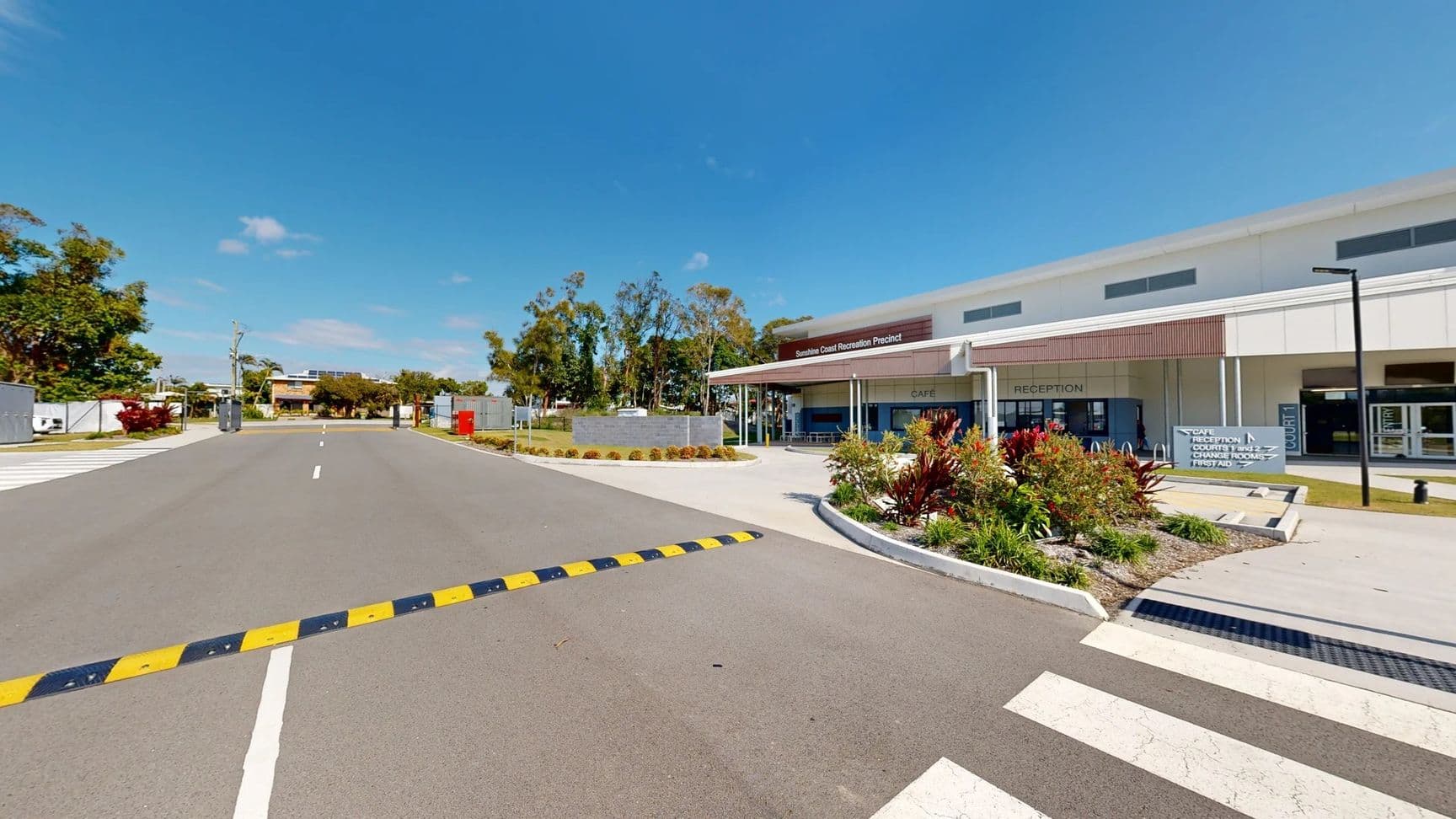 A wide grey asphalt road extends towards a building in the centre. On the left, a landscaped area features various plants and a path leading to an entrance. A red postal box stands prominently. To the right, the modern structure has large glass windows, with visible signage for a café and reception. Brightly coloured foliage outlines the entrance, and a smooth pedestrian crossing indicates safe passage.