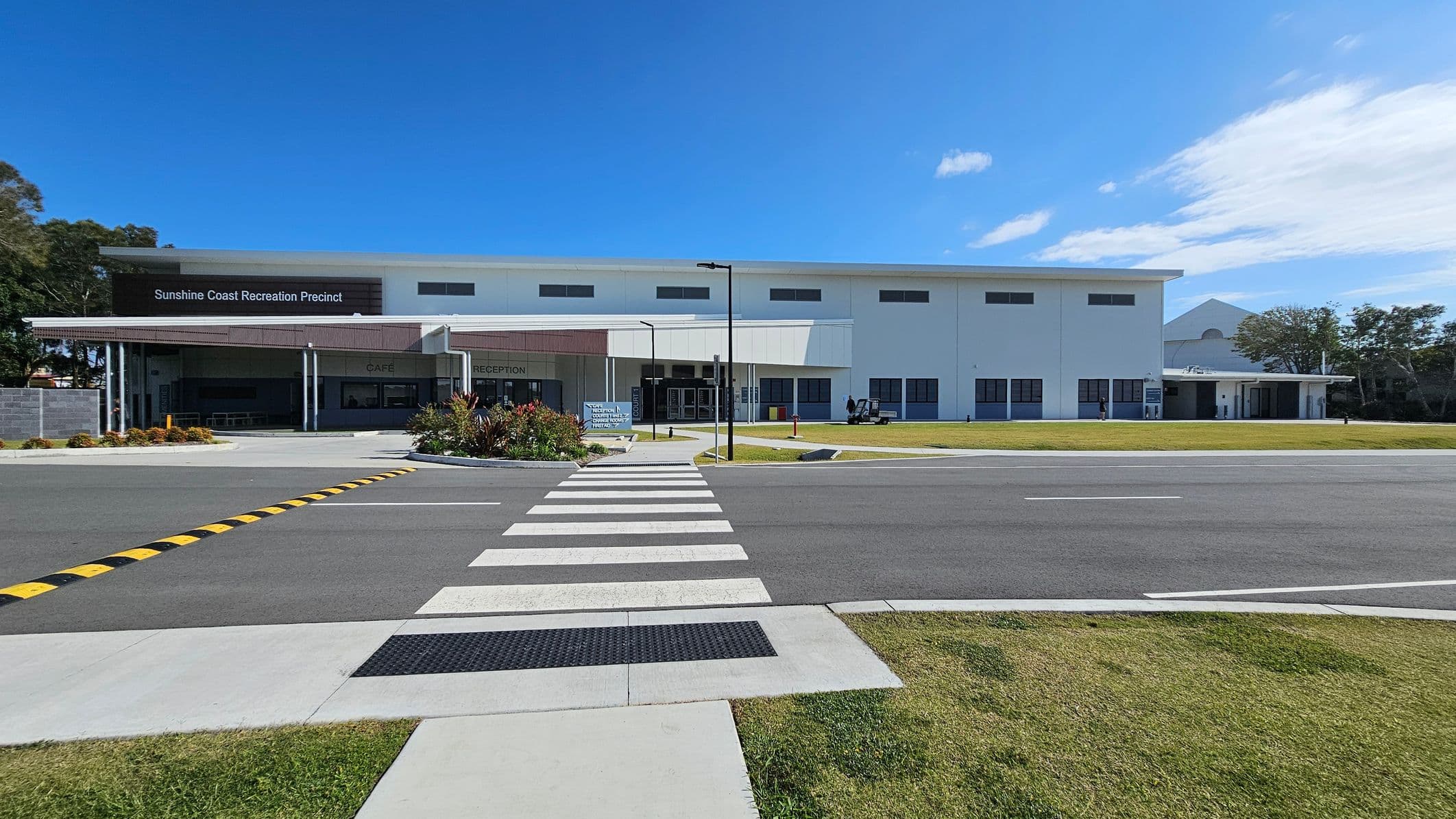 A wide, smooth grey road leads directly to a large white building in the centre, labelled "Sunshine Coast Recreation Precinct." On the left, there is a covered area with outdoor seating and vibrant landscaping. To the right, additional sections of the building include a reception area and an entrance with automatic doors. The foreground features a wide, marked pedestrian crossing with textured pavers, while the background includes patches of grass and trees bordering the area.