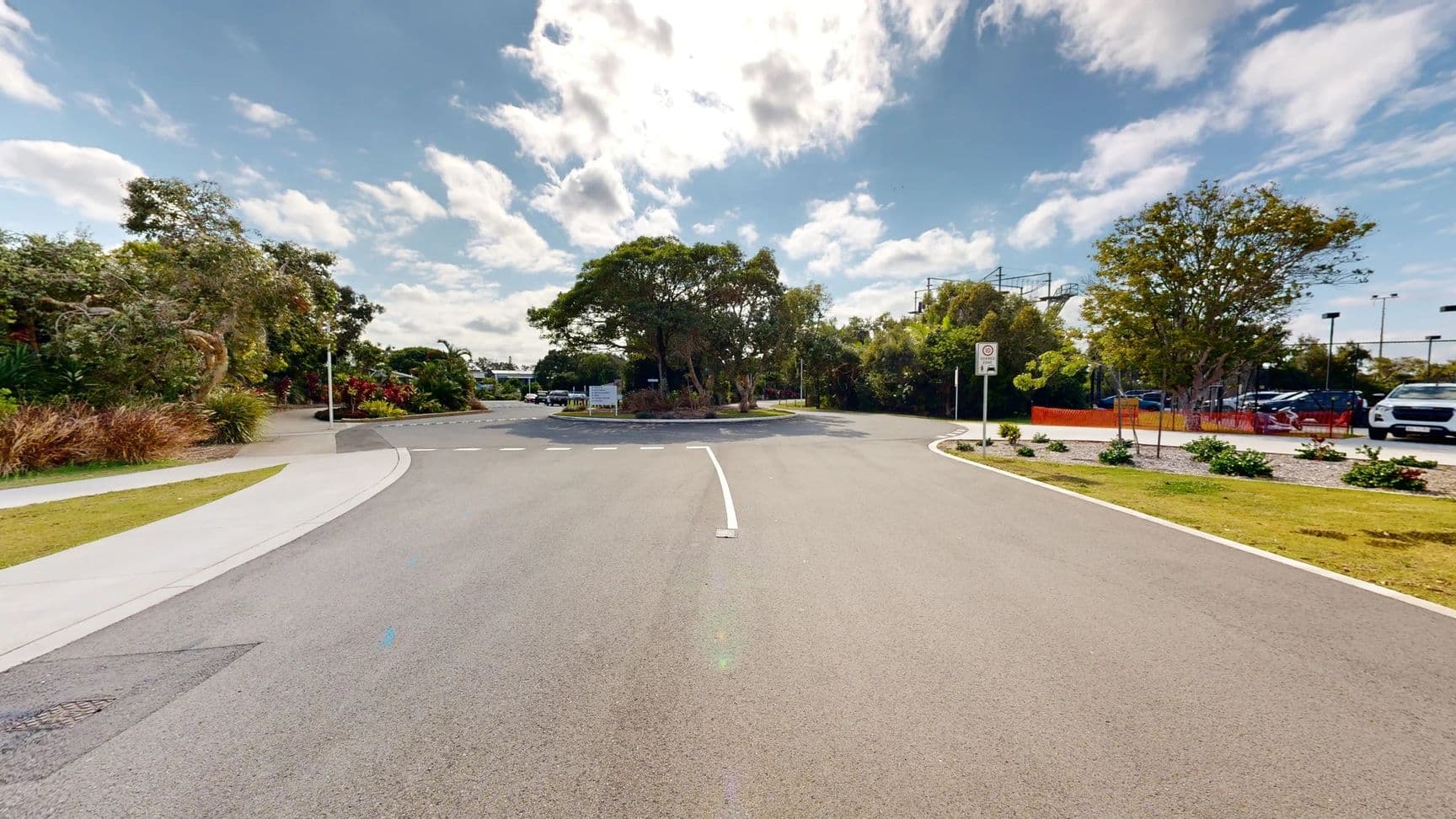 A smooth road curves gently ahead, leading into a roundabout. On the left, a well-kept landscape features various shrubs and small trees, with a paved walkway alongside. To the right, there is a parking area, partially obscured by greenery, and several vehicles are parked in view. In the foreground, the clean road surface is clearly marked with a white line indicating the curve. The background includes more dense foliage, with a large tree providing shade near the centre of the roundabout.
