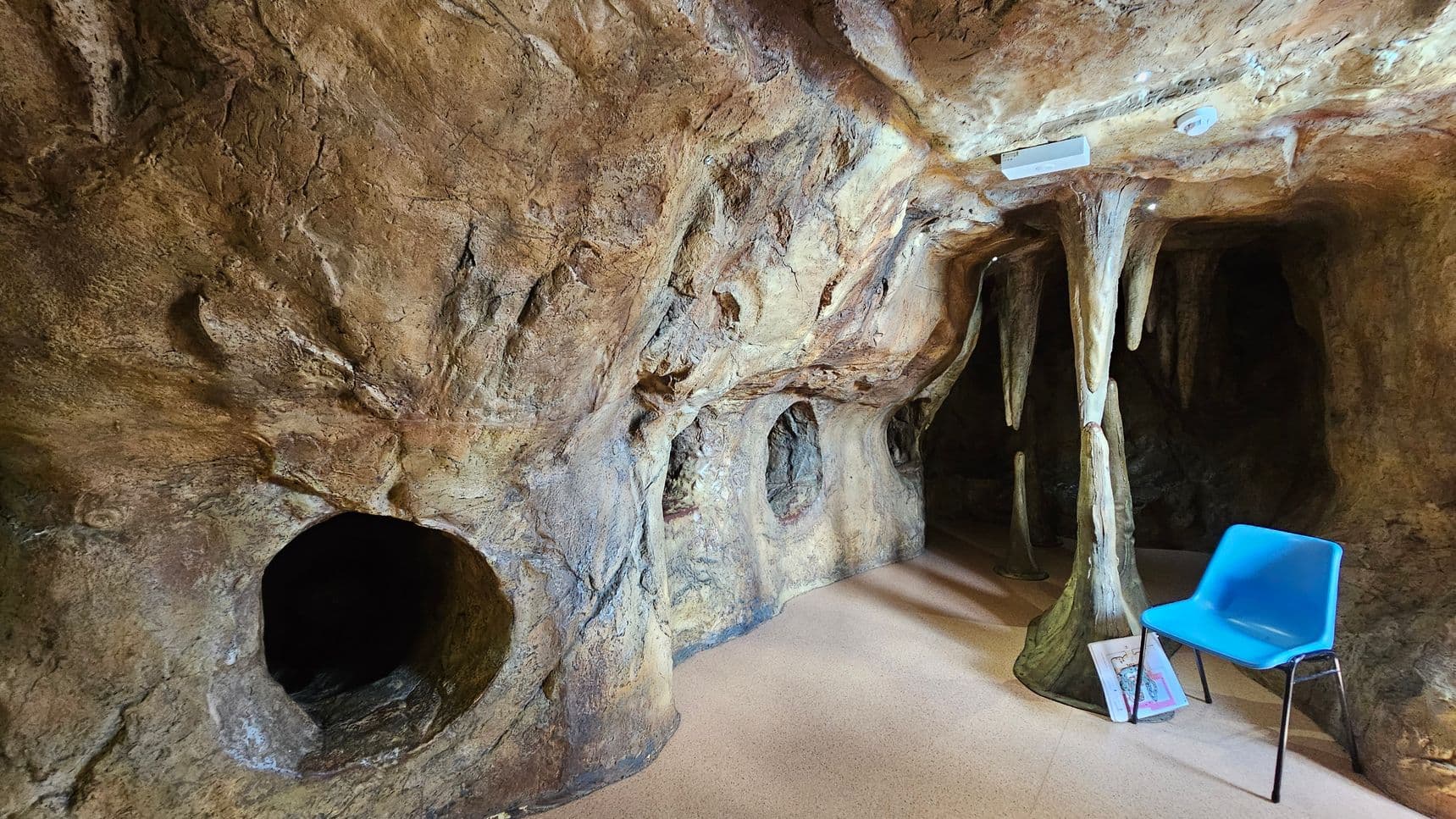 A cave-like room with textured, rocky walls and ceiling. On the right, a blue plastic chair sits on a beige floor, next to a stack of books. Stalactite and stalagmite formations are present, with one column reaching from floor to ceiling. The left side features circular openings in the rock wall, leading to dark recesses. The lighting is dim, enhancing the cave atmosphere.