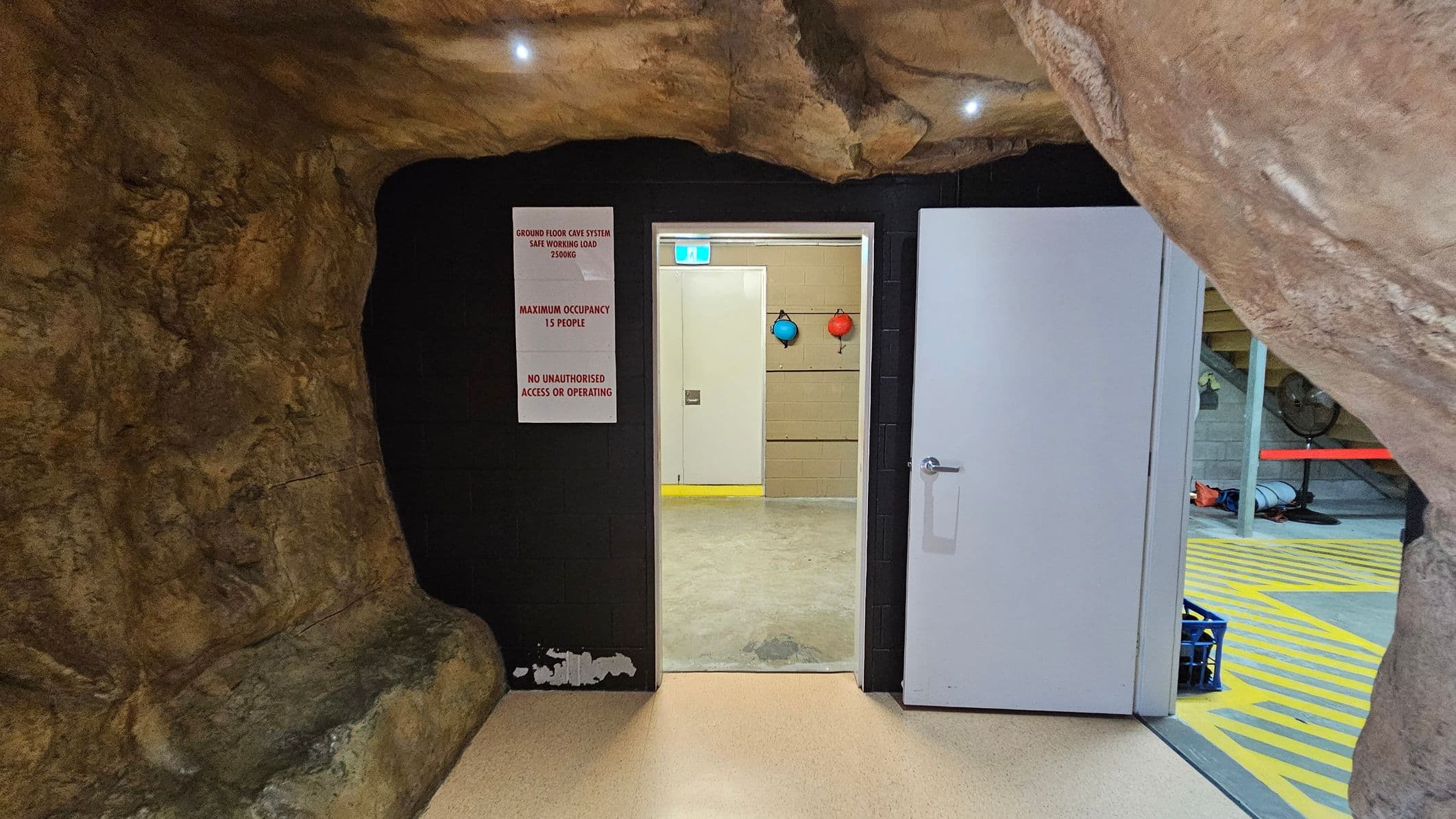 A cave-like interior with rough, rocky walls and ceiling. In the centre, a white door is open, leading to a room with a concrete floor and beige walls. Two coloured buoys hang on the wall inside. A sign on the left of the door reads "Ground Floor Cave System Safe Working Load 1500kg, Maximum Occupancy 15 People, No Unauthorised Access or Operating." The floor is light-coloured, and the ceiling has small recessed lights.