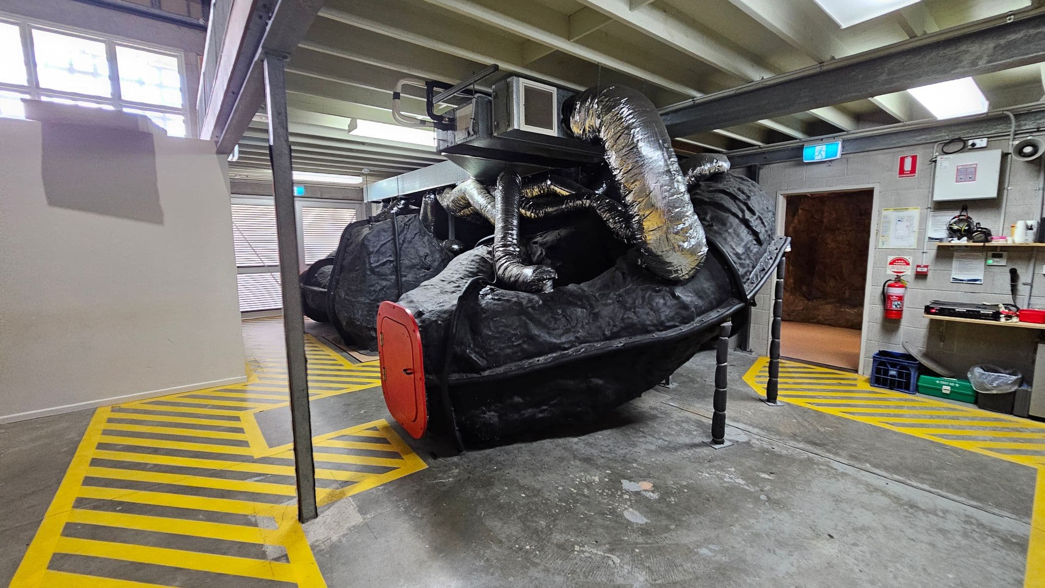 A large, black, rock-like structure with a red door is in the centre, supported by metal legs. Silver ducting tubes are attached to the top, connected to a ventilation unit. The floor has yellow and black striped markings. On the right, there's a wall with a fire extinguisher, safety signs, and shelves holding various items. The ceiling is made of exposed beams with fluorescent lights. Windows on the left let in natural light.