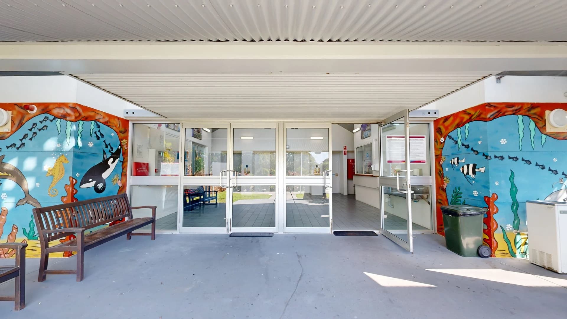On the left, a sign provides information about the facility, mounted on a white pole, with a colourful mural of sea life visible beside it. To the right, a large rock with a sign labelled “Caves Rock Face” sits near vibrant tropical plants. The pathway is made of smooth grey concrete, leading towards double glass doors. The ceiling above is gently sloped, with a large overhang sheltering the entrance. The surrounding area features well-maintained grass and various tropical foliage.