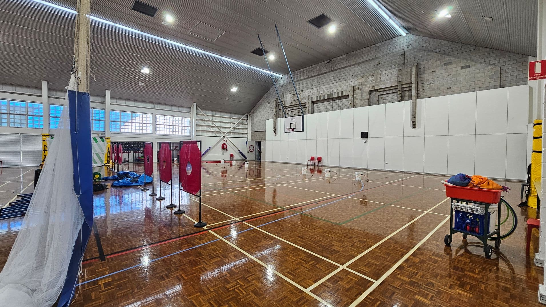 On the left, a tall blue structure is partially covered by a net, with various sports equipment scattered nearby, including a blue tarp. Red targets on stands are lined up in the centre. To the right, a white wall features a basketball hoop, with two red chairs placed beneath it. The floor is polished wood with visible lines indicating court boundaries. The ceiling is high and features a textured design, with lights evenly distributed across its surface.