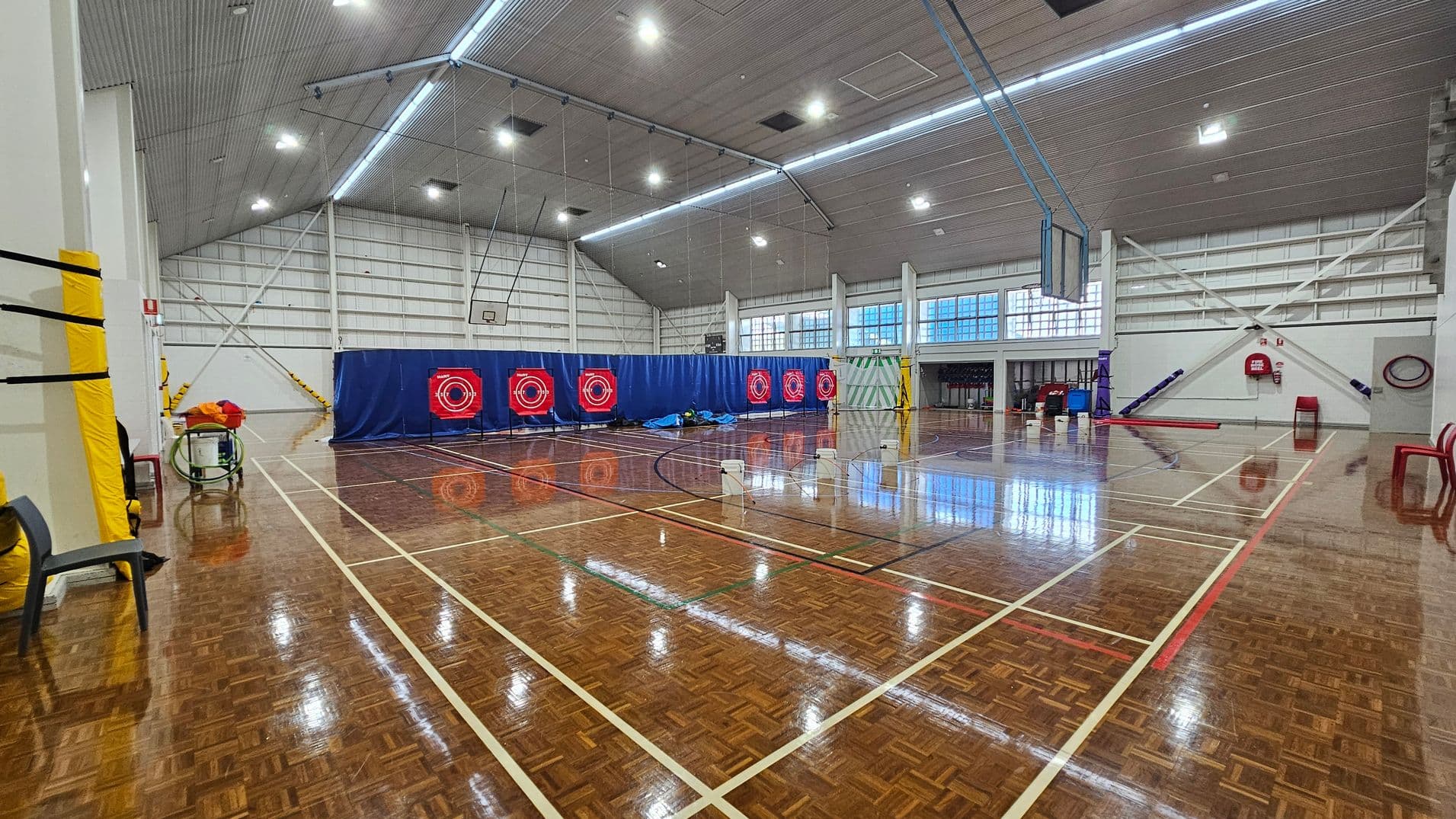 On the left, a tall wall has basketball hoops attached, with a red padded area below. Nearby are various colourful mats stacked against the wall, providing cushioning for activities. To the right, a scoreboard is mounted, along with large windows that allow natural light into the space. The polished wooden floor features court markings in white and green, extending across the area. The ceiling is sloped with bright lighting evenly distributed to illuminate the gymnasium effectively.