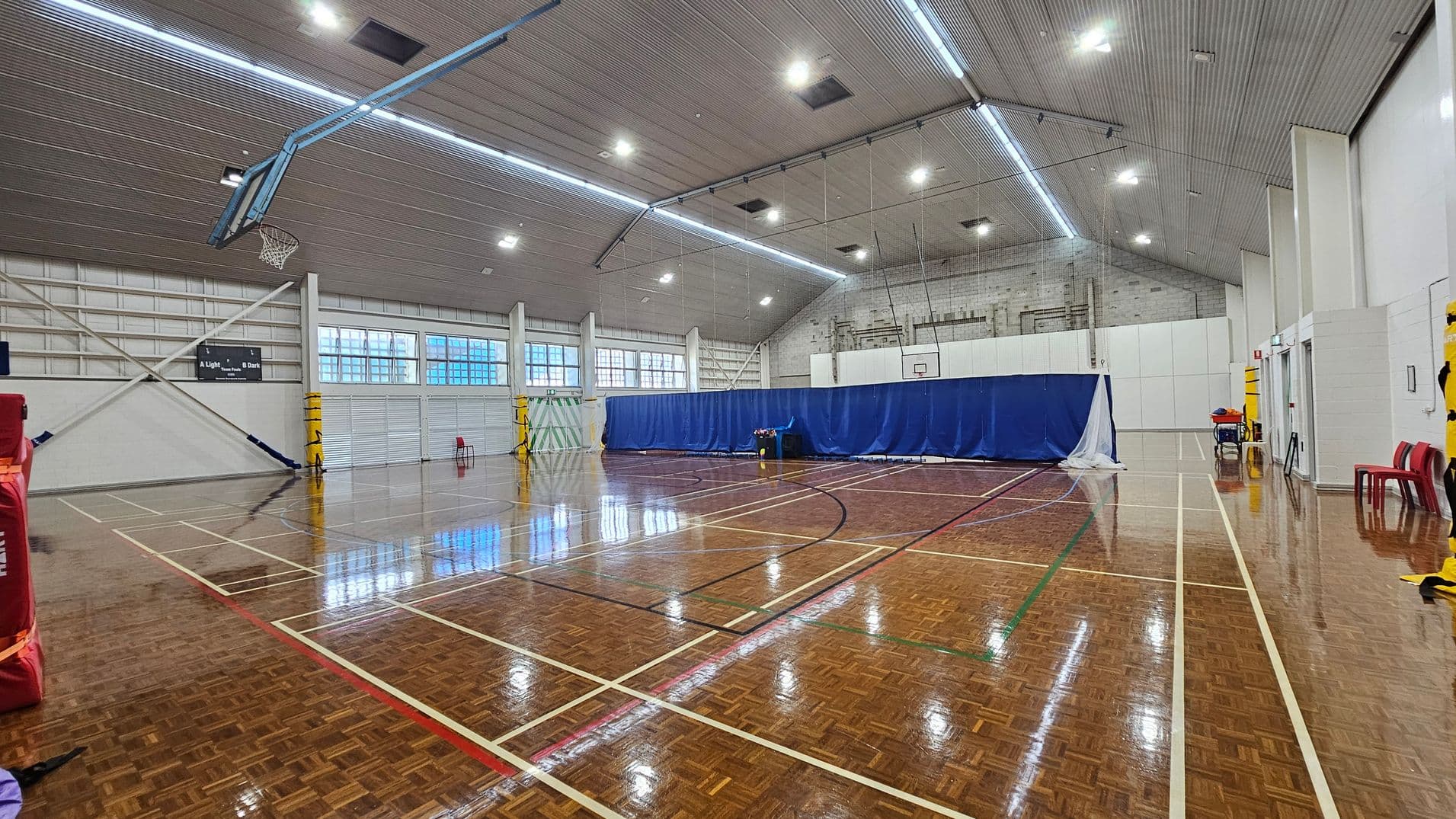 On the left, a red padded wall is partially visible, with a basketball hoop fixed above it, and large windows allowing light into the space. To the right, a blue curtain separates part of the area, with a couple of red chairs positioned nearby. The glossy wooden floor features white and coloured lines marking different court boundaries. The ceiling is high with a ribbed texture, fitted with bright overhead lights that provide ample illumination throughout the gymnasium.