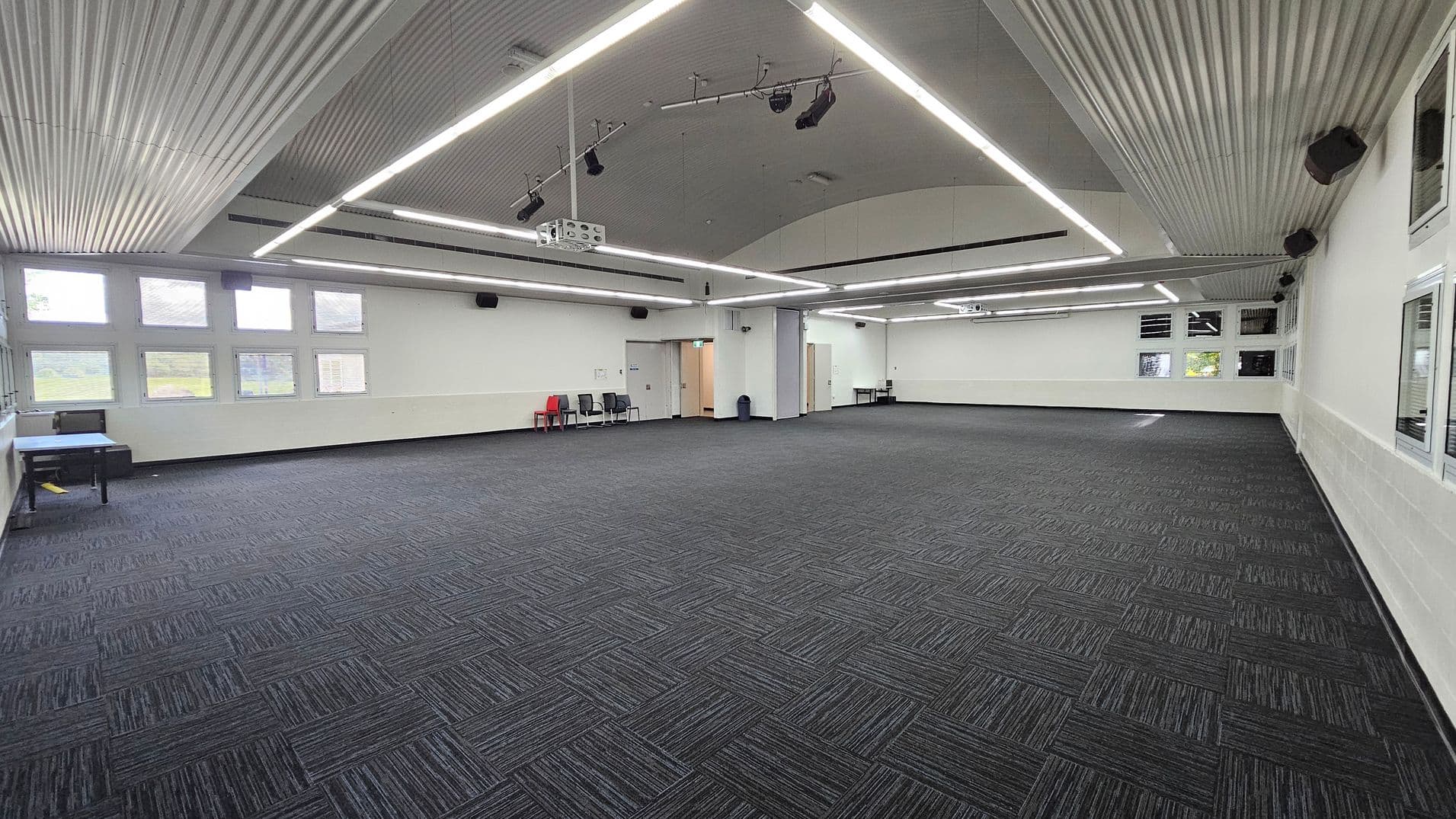 On the left, a small table with a blue cloth is set against the wall, accompanied by a few grey chairs. To the right, a doorway leads to another room, and there are additional chairs positioned nearby. The floor is carpeted in a dark pattern, and the spacious area is largely empty. The ceiling is curved with a corrugated design, featuring bright LED lights running across it, providing even illumination throughout the room.