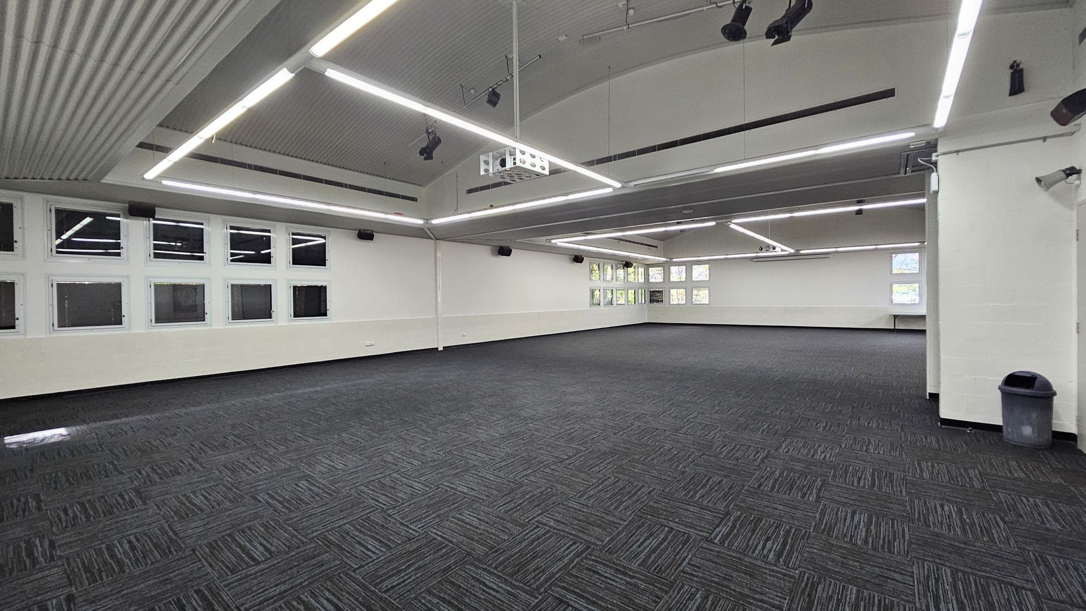 On the left, a row of small windows is situated just above a low white wall, allowing light to enter the room. To the right, a grey rubbish bin is positioned next to a small corner area with a low countertop that is unobtrusive. The floor is covered in dark, textured carpeting laid in a patterned design. The ceiling arches gently and features several bright, linear lights along its length, illuminating the spacious interior.