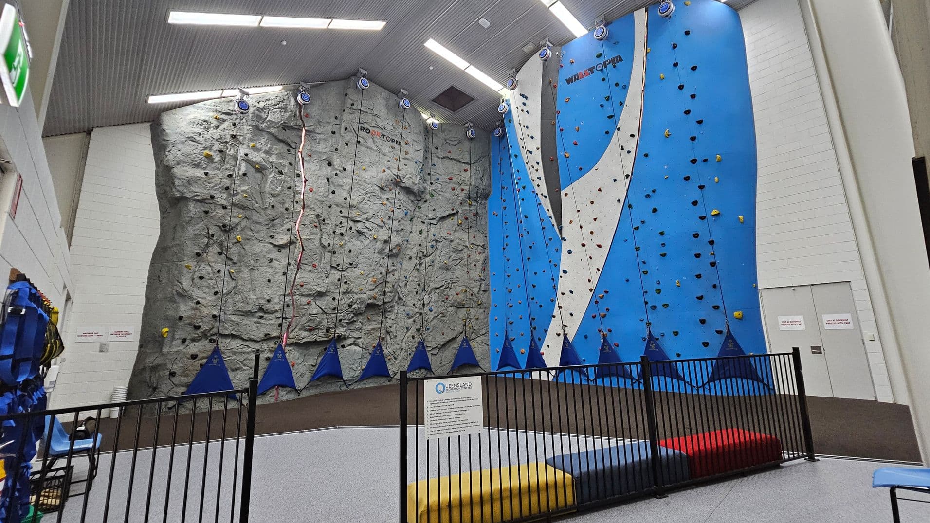 Double doors are open, revealing a soft, grey floor and a low wall with a safety railing on the left. A vibrant red mat is positioned in the centre, with blue climbing shapes in the background, hinting at climbing activities. To the right, there's a wall decorated with the words "The Rock Face" in bold, modern typography, accompanied by a geometric blue design. The ceiling is plain and light-coloured, providing a subtle contrast to the walls.