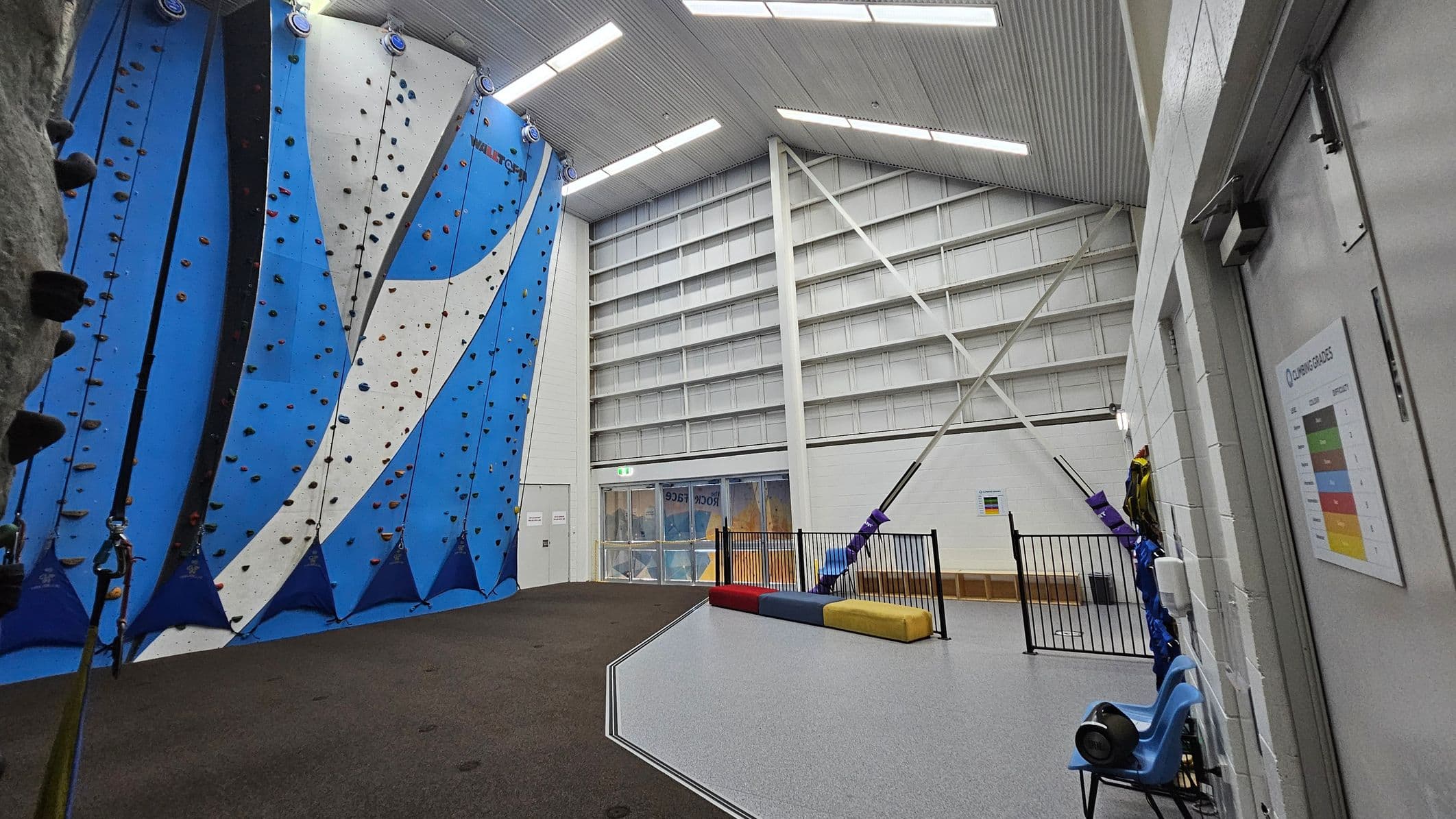 Indoor climbing gym with blue and white climbing walls on the left, featuring various holds. The floor is a mix of dark and light grey surfaces. On the right, a white wall with a climbing grades chart and a blue chair. In the background, a large windowed exit door and a fenced-off area with colourful padded blocks. The ceiling is high with exposed beams and fluorescent lights.