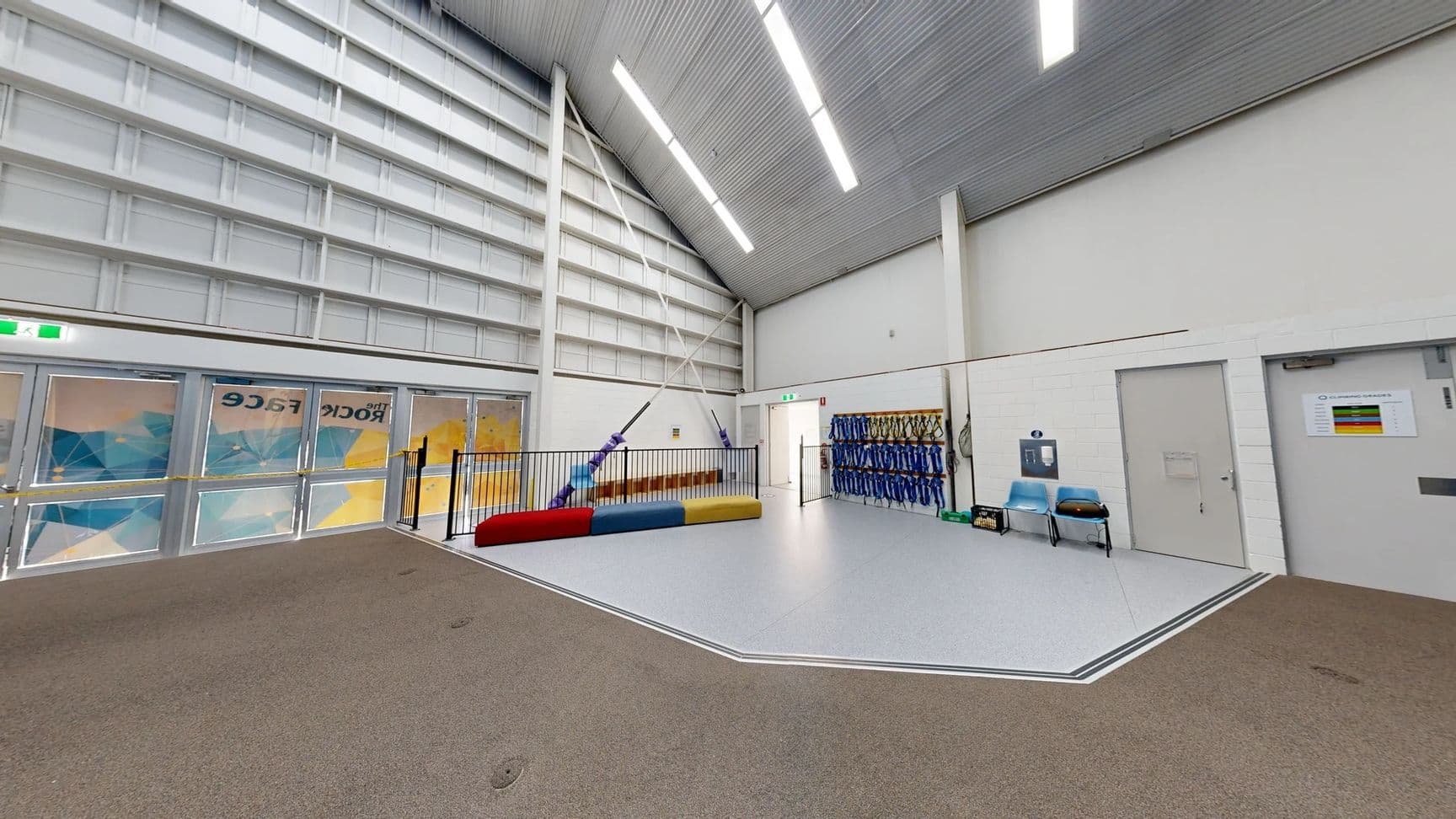On the left, large double doors with patterned glass reveal a vibrant, geometric design behind them. A railing surrounds a colourful mat in red, yellow, and blue, positioned in the centre of the space. To the right, a wall displays several rows of blue harnesses, and two light blue chairs are placed nearby. The floor has a mix of light grey and soft brown textures. The ceiling is high with a series of linear lights running centrally, and is lined with corrugated metal.