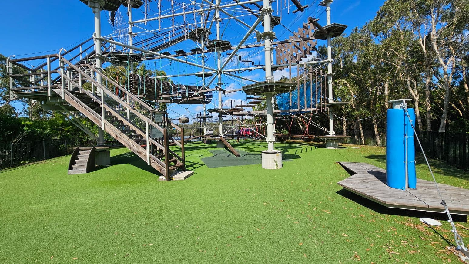 On the left, there are metal bleachers with a white canopy above, supported by metal poles. In the centre, a large outdoor climbing structure with ropes and platforms is visible, labelled "Sunshine Coast." The ground is paved with concrete, leading to a grassy area. On the right, there are various green and red-leaved plants and trees, providing a natural backdrop.