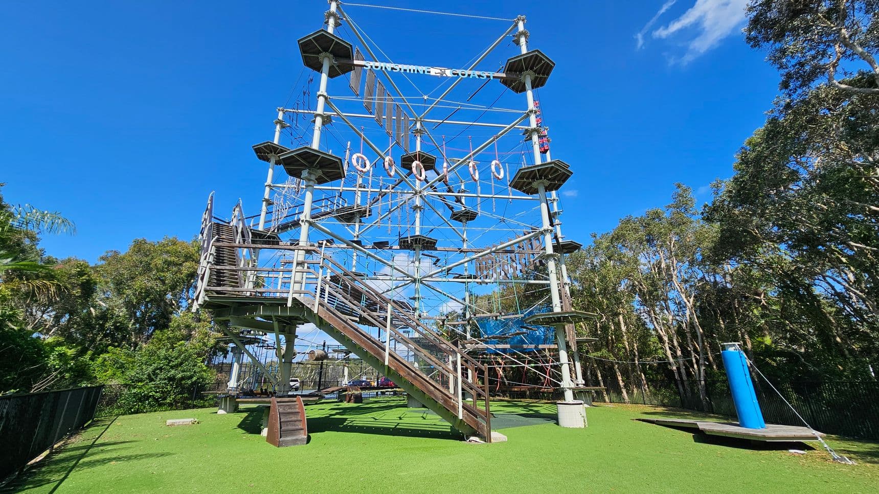 A large outdoor climbing structure with multiple levels and platforms, featuring ropes and obstacles. The structure is supported by metal poles and has a staircase on the left side. The ground is covered with artificial green grass. Surrounding the structure are tall trees and a clear blue sky. A blue cylindrical object is on the right side near the base.