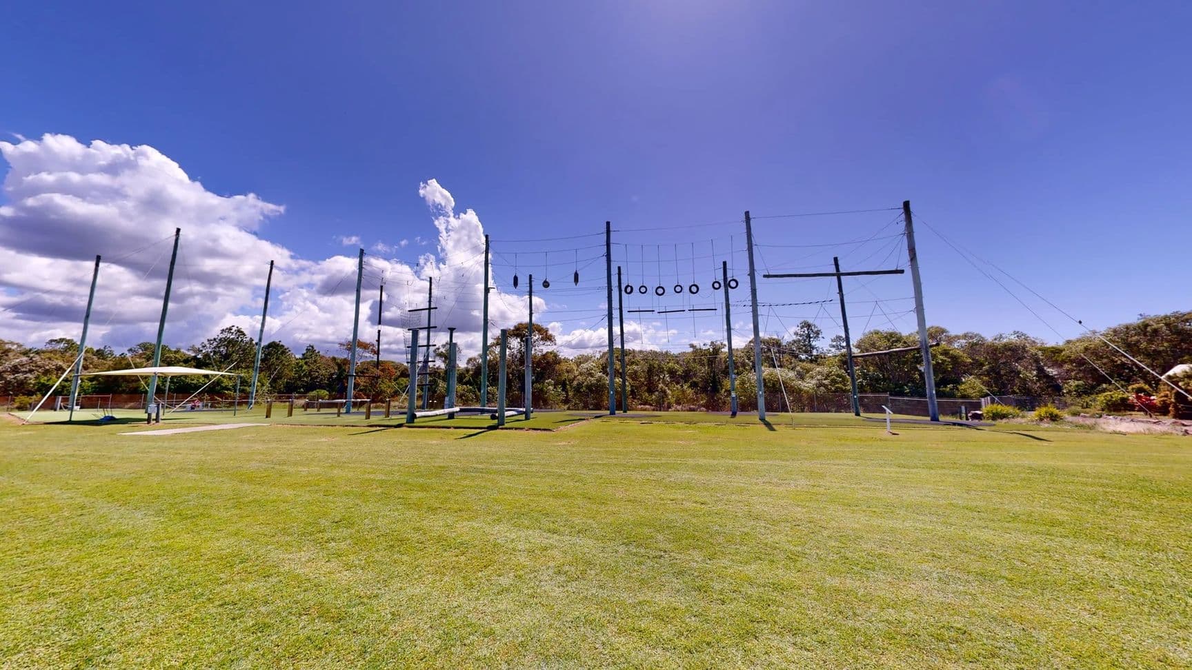 To the left, there is a large, green, rectangular area of grass, alongside a low structure that appears to be a support for a zip line or swinging apparatus. To the right, several vertical poles are arranged, some with horizontal bars and others adorned with tyres, indicating a challenge course. In the background, lush trees provide coverage, while the foreground is a well-kept expanse of grass. No notable details of the floor or ceiling are visible in this setting.