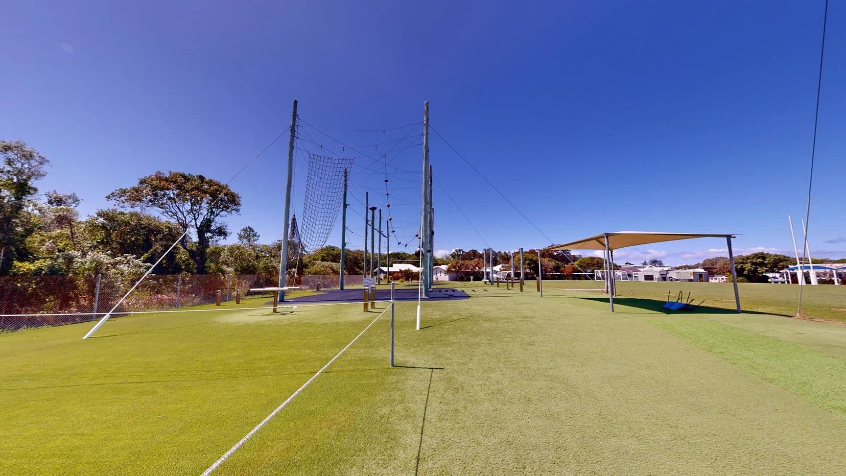 On the left, a series of tall poles are set up, connected by wires and nets, forming an obstacle course. A dark rubber flooring area is visible beneath them. On the right, there is a shaded structure with a sloping roof, supported by poles, and a blue mat or equipment situated beneath it. The ground is covered in even green artificial grass, extending across the scene. In the background, trees and low buildings are present, providing a natural setting.