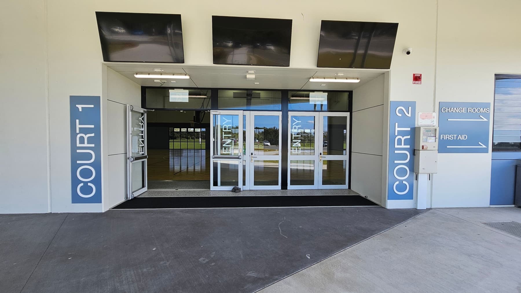 At the entrance, two sets of glass doors lead into a spacious interior. On the left, a large panel displays "COURT 1," while on the right, another panel indicates "COURT 2," alongside directional signs for "CHANGE ROOMS" and "FIRST AID." Above the doors, there are three dark ceiling panels, and fluorescent lights illuminate the entrance area. The ground is a smooth concrete surface with a slight slope towards the entrance, providing easy access.