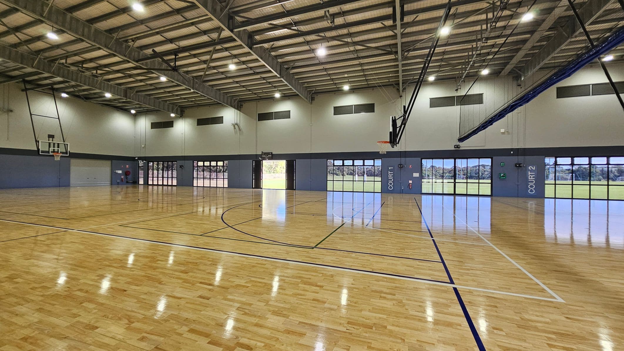 In a large indoor sports hall, polished wooden floors stretch across the area, reflecting the light from overhead fluorescent fixtures. On the left side, a basketball hoop hangs above a blue wall with an entrance door nearby. To the right, two large glass doors marked "COURT 2" reveal an outdoor field beyond. Each side features large windows allowing natural light into the space. The ceiling is high and lined with metal beams, creating an open atmosphere.