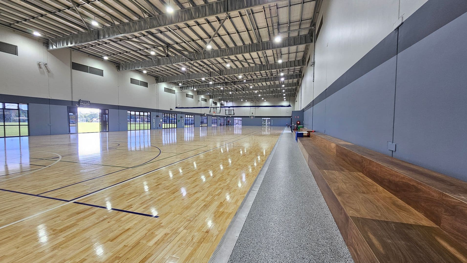 The sports hall features polished wooden flooring that reflects overhead lights, with court markings visible across the surface. On the left, a smooth concrete area runs parallel to the court, leading to a wooden bench that provides seating. To the right, a blue wall has a door and safety equipment visible, including a fire extinguisher. The ceiling is high, constructed with metal beams and lined with bright fluorescent lights that illuminate the space evenly.