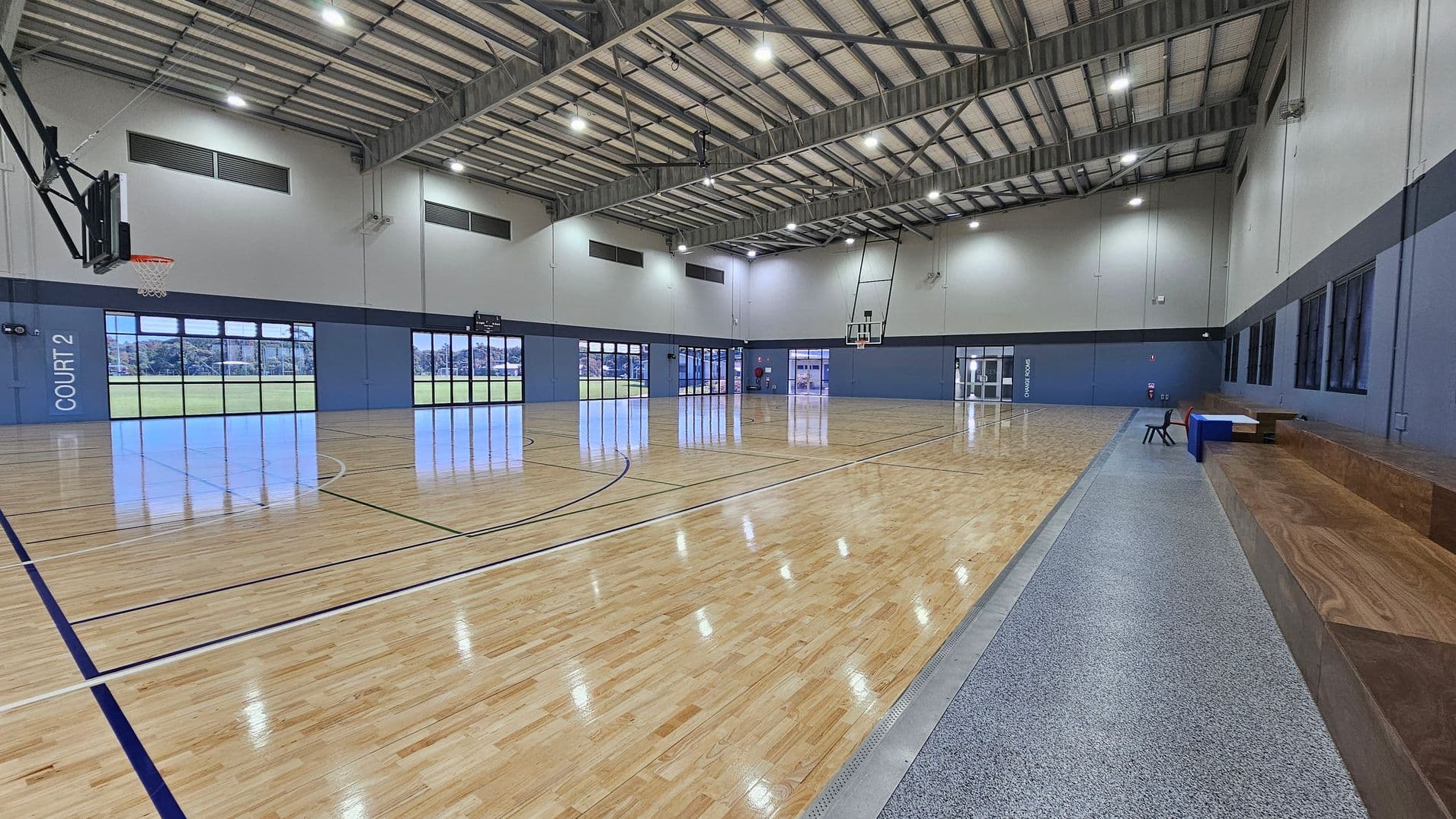 In a spacious sports hall, polished wooden flooring extends across the area, marked with blue and white lines for court play. On the left side, a basketball hoop is mounted above a grey wall, while "COURT 2" is clearly marked beneath large windows that provide views of the outside. To the right, glass doors lead to an entrance, and a raised wooden seating area runs along the wall, providing a place for spectators. The ceiling is high with exposed metal beams and bright overhead lights.
