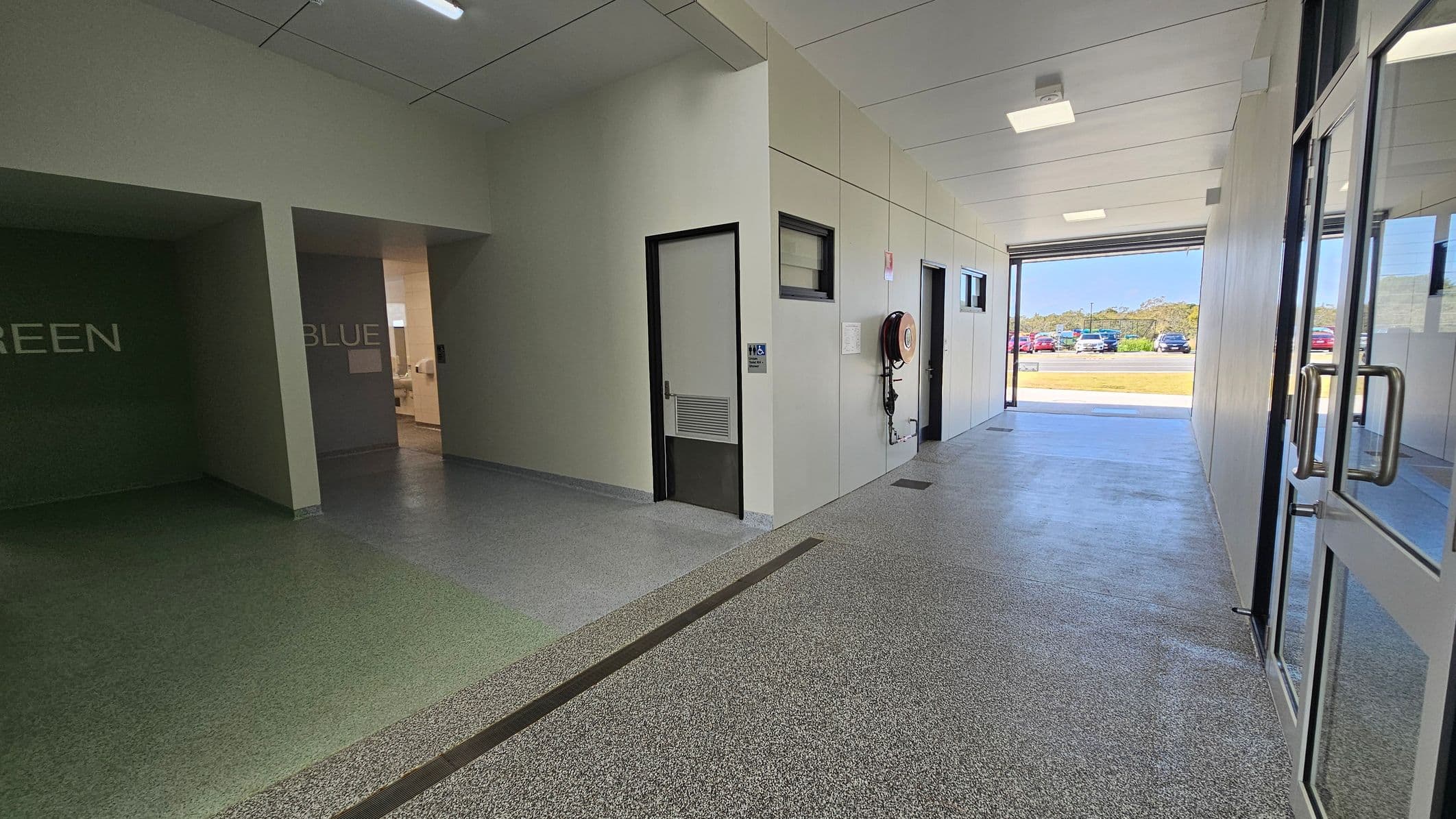 In a corridor, the floor features a textured combination of green and grey surfaces, smoothly transitioning towards a polished concrete section at the far end. On the left, a wall painted in a muted green displays the word "GREEN," with an adjoining door leading to another room. To the right, another wall marked with "BLUE" has a door beside it, which opens into a space with additional facilities. The ceiling is flat and white, fitted with square fluorescent lights.