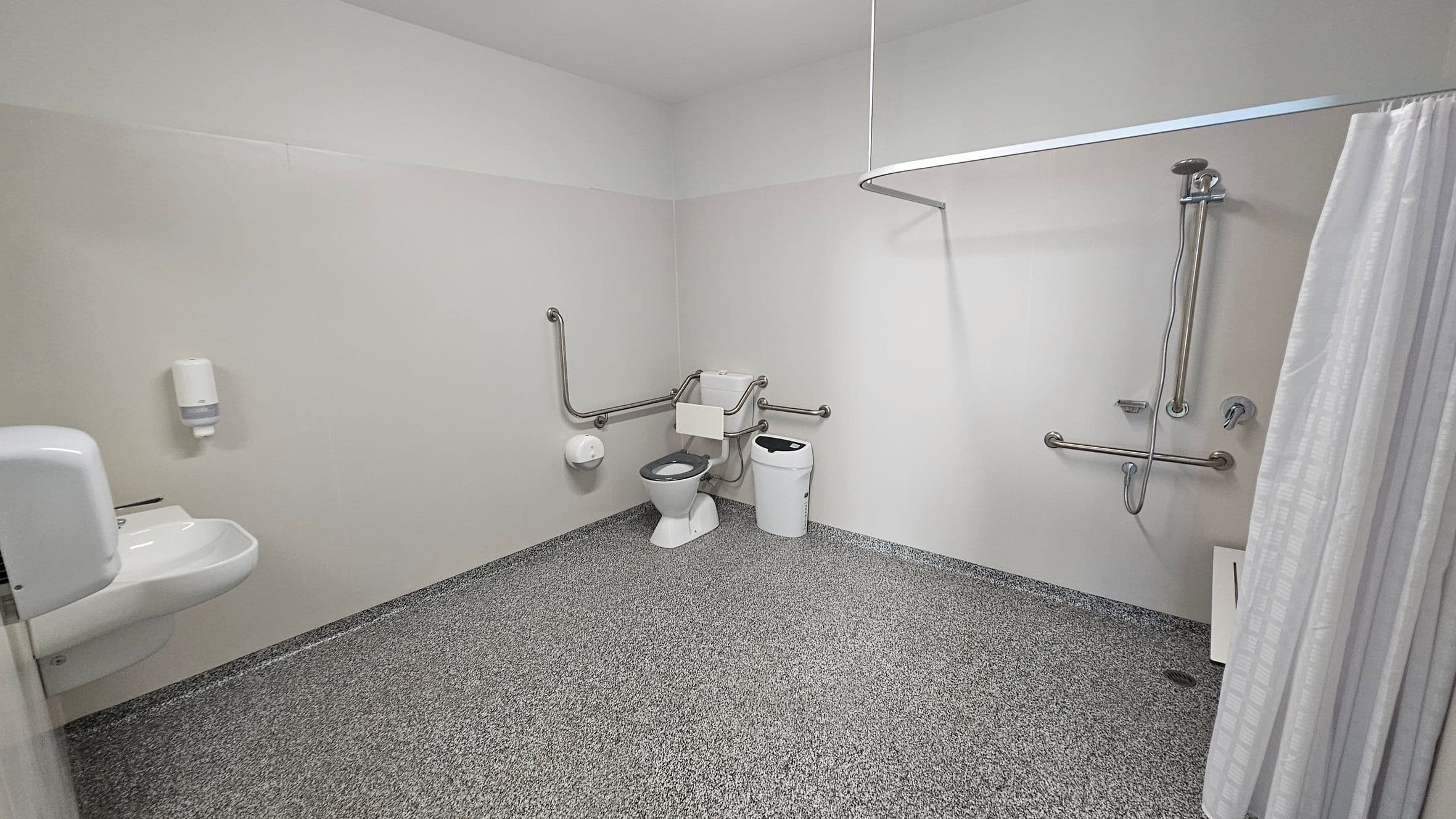 The room features a textured grey floor that extends throughout the space, providing a non-slip surface. On the left, a white sink is mounted on the wall with a soap dispenser above it. Adjacent to this is a toilet, equipped with grab bars for accessibility. On the right side, a shower area is enclosed by a curtain, with additional grab bars mounted on the wall next to the shower head. A small, rectangular waste bin sits beside the toilet, and the ceiling is flat and finished in white.