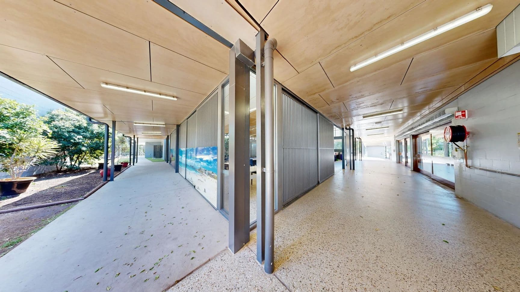 On the left, a sloped pathway leads towards a smooth concrete surface bordered by a low wall. There’s a white door with a metal handle and a small sign next to it. Mounted above, there are multiple fire alarms and a wall sign with safety information. On the right, a series of glass panels reveal a corridor with additional doors further down. The floor is a light grey concrete, and the ceiling is flat, featuring wooden panels with fluorescent lighting fixtures spaced evenly.