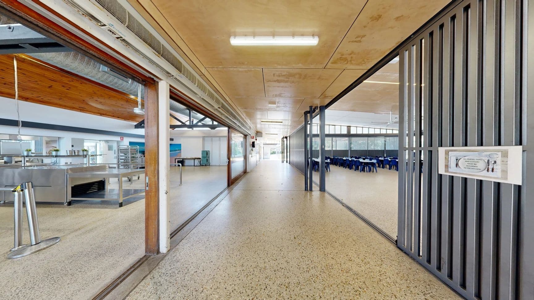 On the left, there is a wide entrance leading into a kitchen area with stainless steel equipment and shelving visible, along with a light-coloured wall. A smooth, polished floor extends throughout. On the right, a grey partition separates the corridor from a dining area filled with rows of blue seats around tables. The ceiling features a combination of flat panels and exposed beams, with overhead lights illuminating the space.
