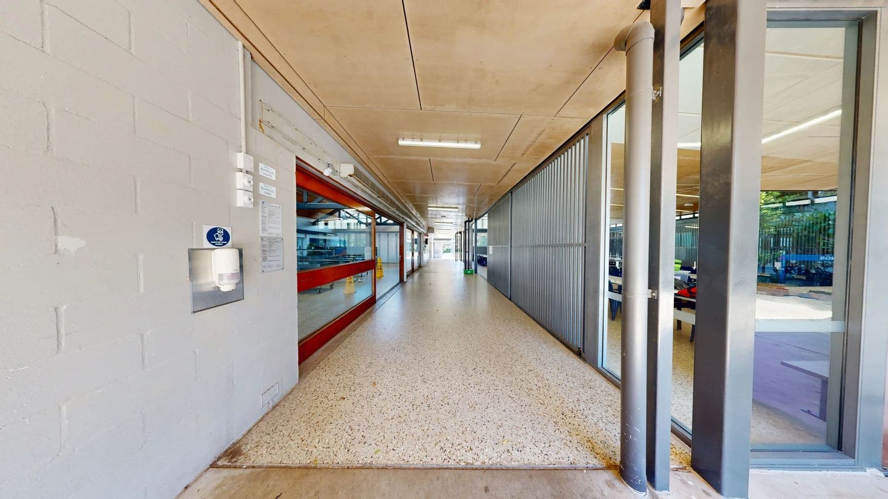 On the left, a light grey concrete wall features a paper towel dispenser and several mounted notices. A red-framed window offers a view into a room with equipment. The floor is a smooth, speckled surface that transitions to wood near the edge. On the right, large glass panels are set in a steel frame, revealing a corridor with blue tables and chairs in the background. The ceiling is a flat concrete design with strip lighting running along its length.