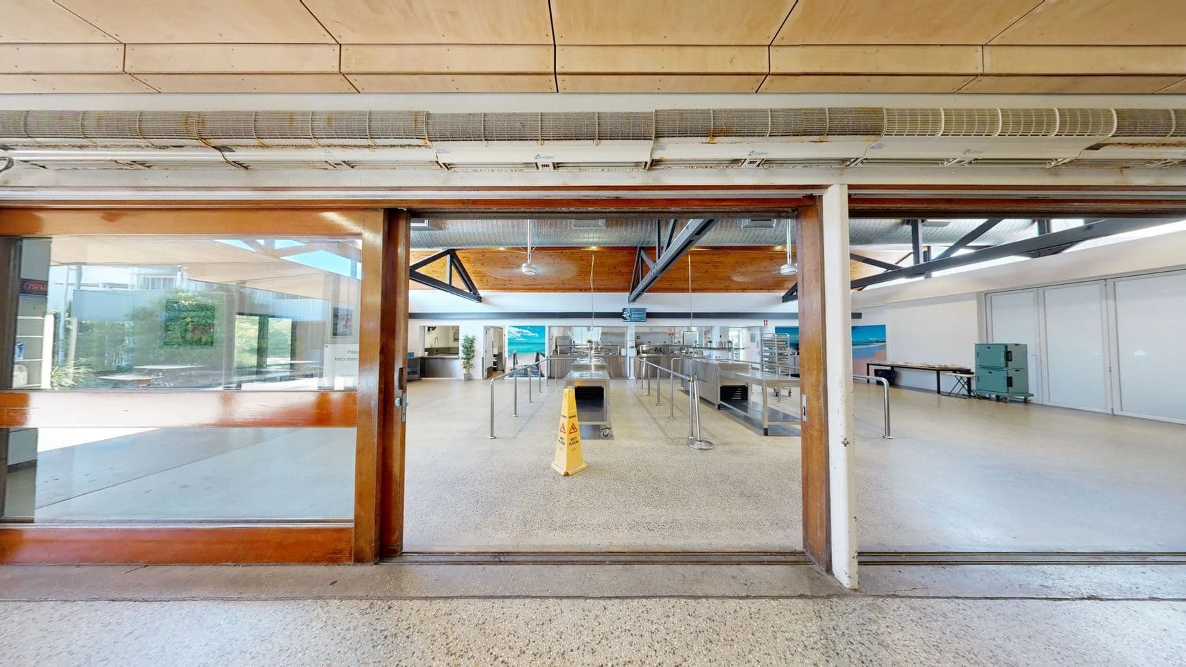 On the left, there’s a large glass door with wooden frames leading to an outdoor area visible through the glass. A small sign is attached to the wall nearby. To the right, a hallway opens into a kitchen area with stainless steel counters and equipment. The floor is smooth, speckled concrete, and the ceiling features wooden panels with exposed beams and overhead lights. In the centre, a caution sign is placed on the floor near a line of barriers guiding visitors towards the service area.