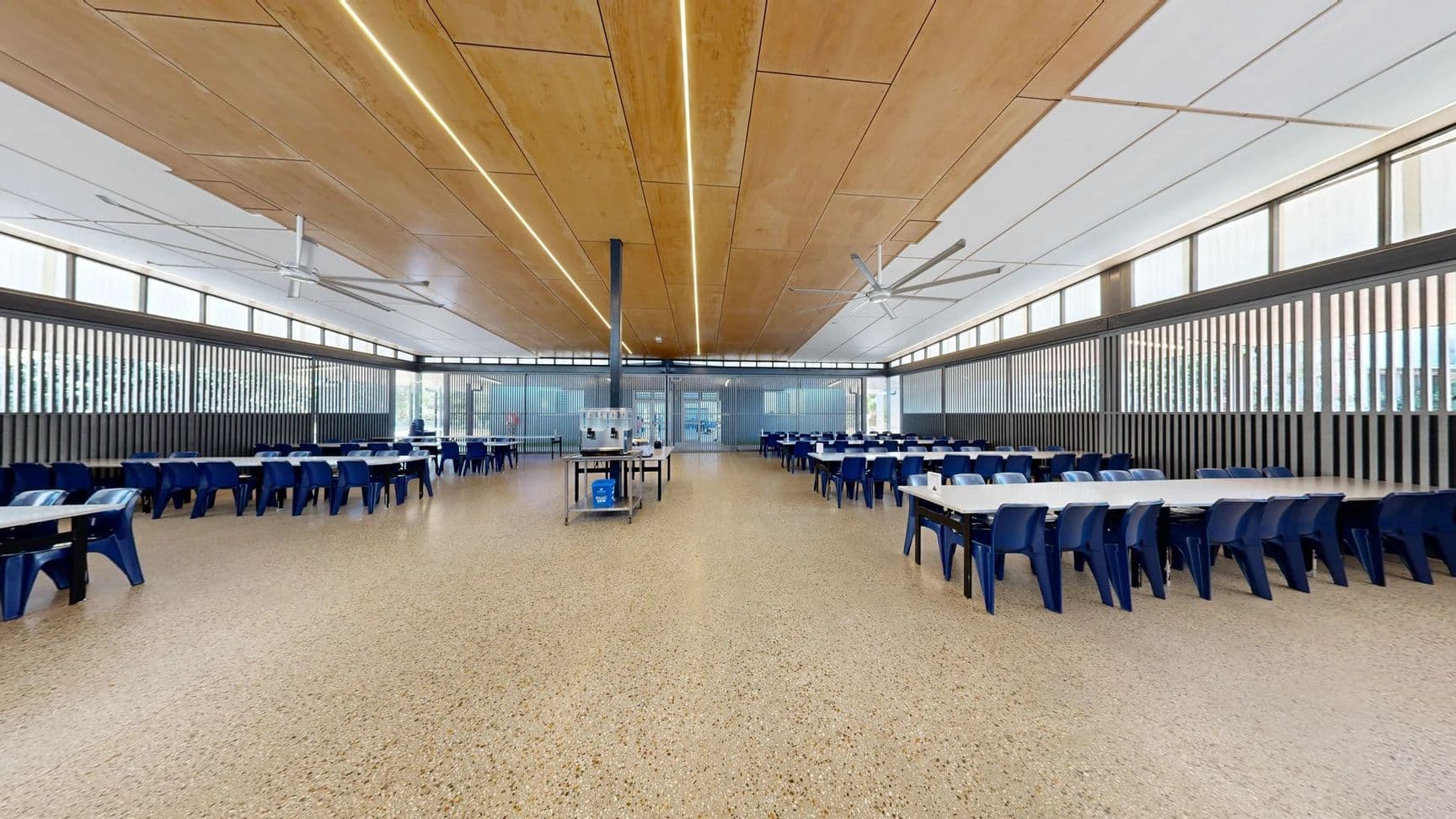 On the left, a row of blue tables and chairs extends along the wall, with additional seating visible further down. On the right, more tables with blue chairs create a symmetrical layout, with narrow vertical windows above. In the centre, a serving cart stands on a polished, speckled concrete floor. The ceiling features wooden panels with integrated lighting and large ceiling fans. The space is well-lit, and the layout is open, with clear pathways between tables leading to a background area.