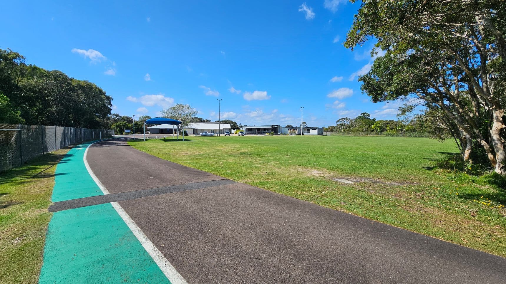 On the left, a narrow path curves alongside a wire fence, with a green strip adjacent to the asphalt surface. The path leads towards a series of buildings in the background, including one with a blue roof. To the right, there is a wide expanse of well-groomed grass. The ground is primarily dark asphalt with a light-coloured border, transitioning to a smooth grassy area.