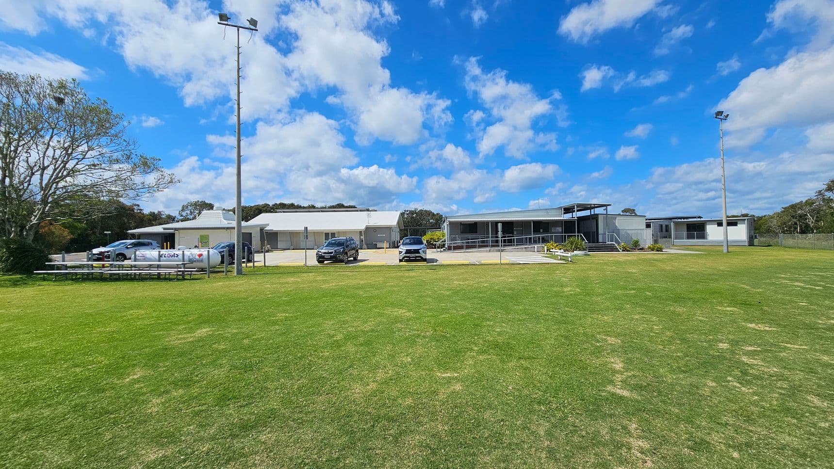 To the left, there are several parked cars near a small structure, with benches lined up parallel to the grass. On the right, a larger building features a raised deck area with steps leading up to it. The floor is a manicured expanse of green grass extending across the foreground, with a light-coloured concrete path visible near the buildings. In the background, there are utility poles and trees providing some greenery around the area. e.