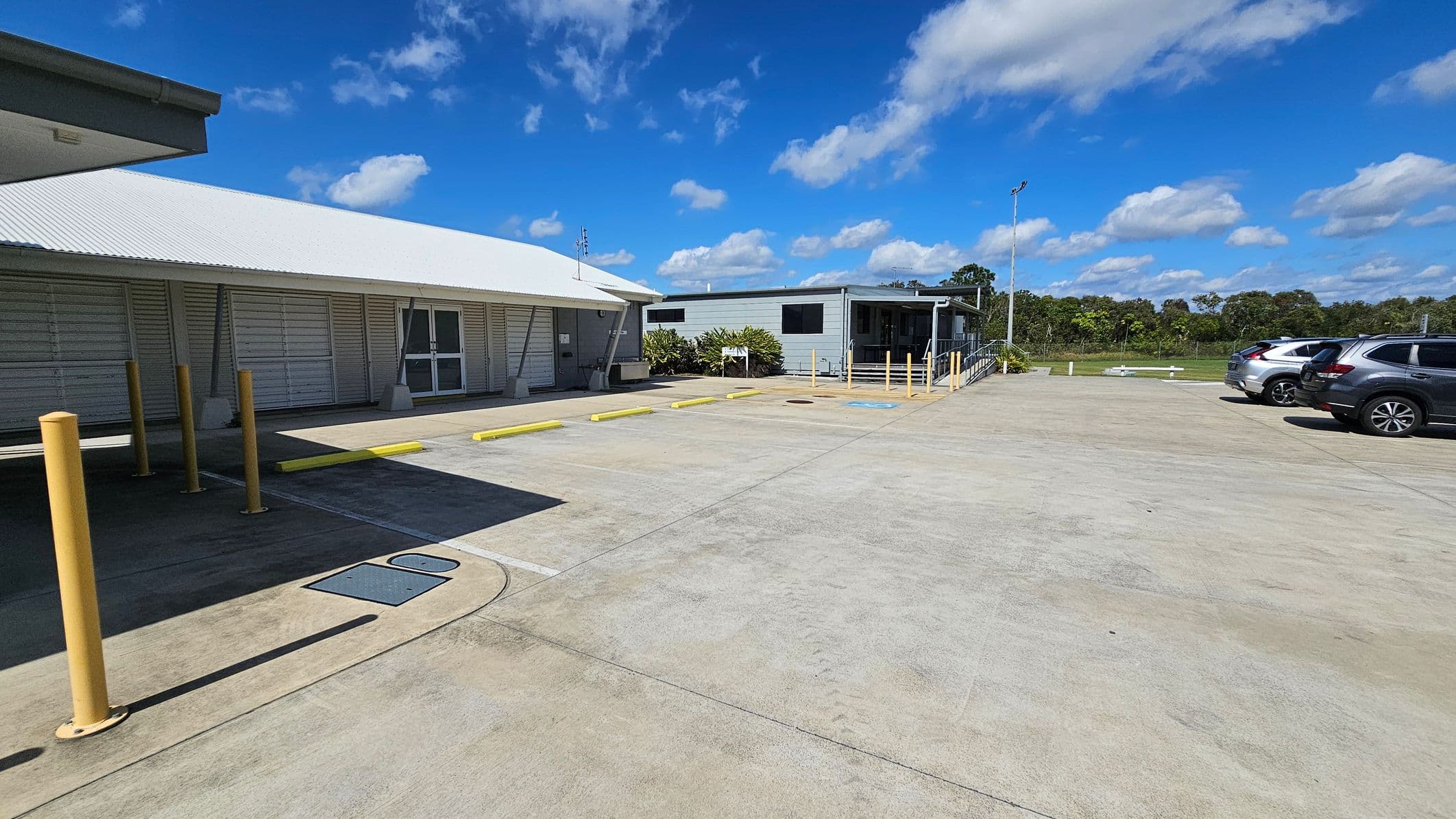 On the left, there is a building with a light-coloured wooden façade and a sloping roof, featuring a set of double doors. In front, yellow bollards line the edge of a concrete parking area. To the right, another structure with a flat roof has a small set of stairs leading up to it, surrounded by some greenery. A few parked cars are visible, occupying a smooth, light grey concrete surface.