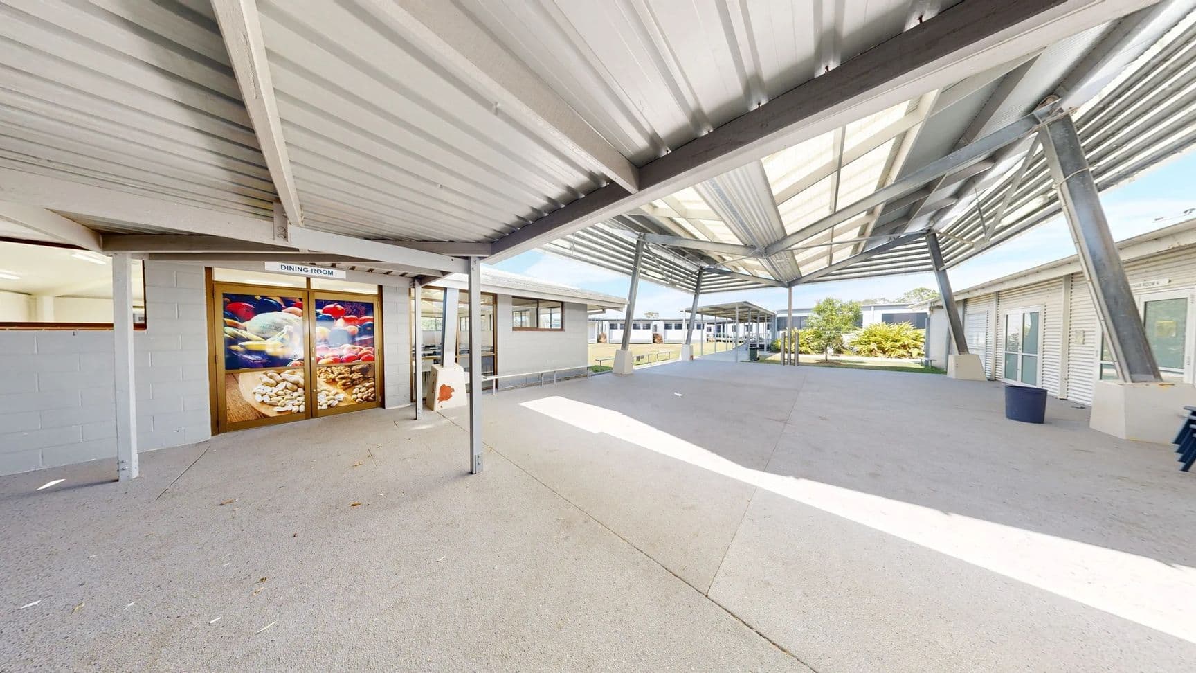 On the left, there is a door marked "DINING ROOM" with a large decorative glass panel featuring colourful fish. Adjacent to it, a grey wall with a window is visible. To the right, additional buildings extend into the background, showing more windows and entrances. The floor is a smooth, pale concrete surface, reflecting light. The ceiling overhead is made of corrugated metal, supported by steel beams, creating a spacious covered area.