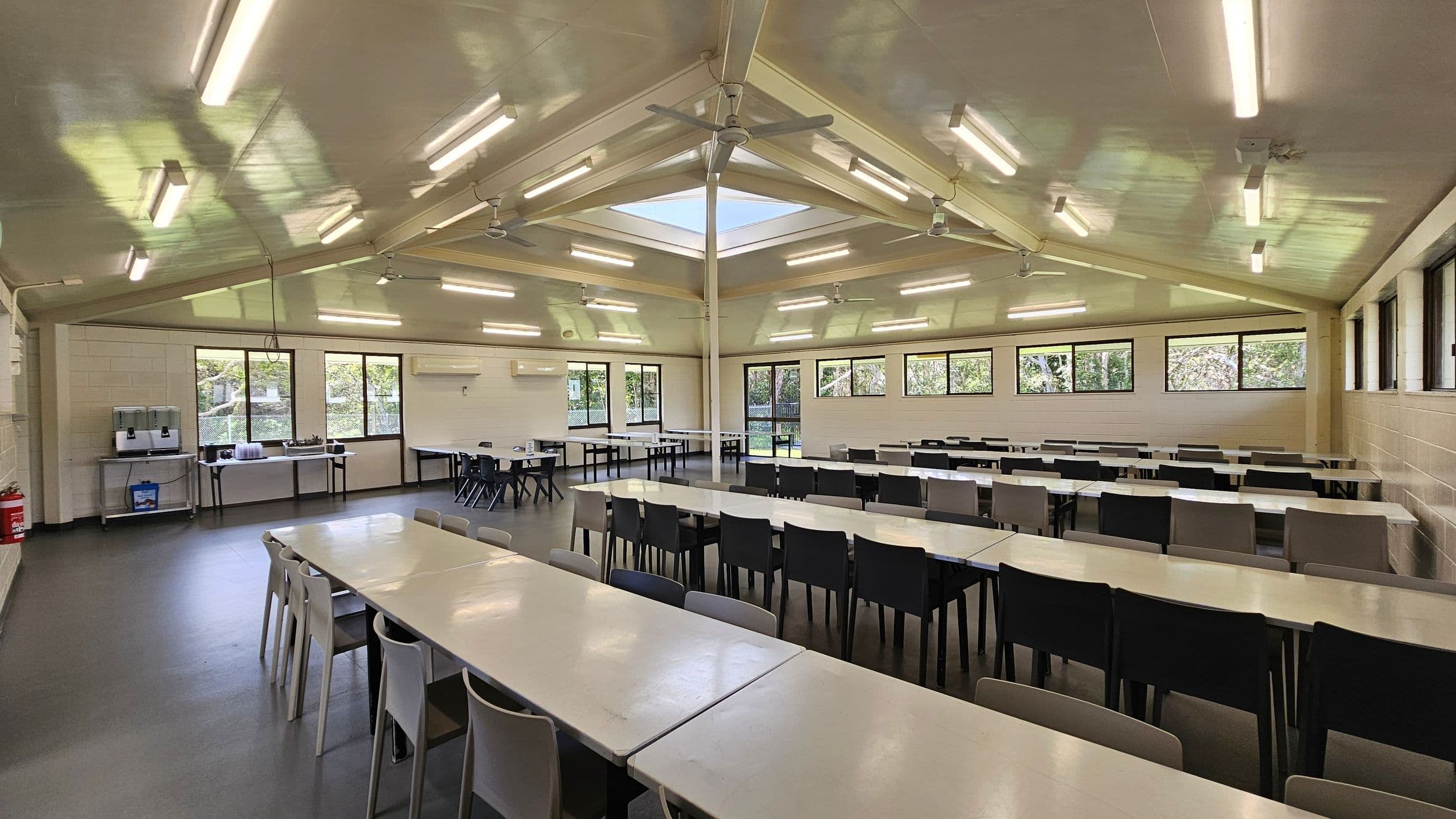 To the left, there are two large windows allowing natural light to enter, with a serving area featuring a water dispenser and trays. Adjacent is a wall-mounted air conditioning unit. On the right, additional windows extend along the wall, providing views of greenery outside. In the middle, multiple long tables with white surfaces are arranged, surrounded by black and grey chairs. The floor is a smooth dark surface, and the ceiling is flat with ceiling fans and fluorescent lights.