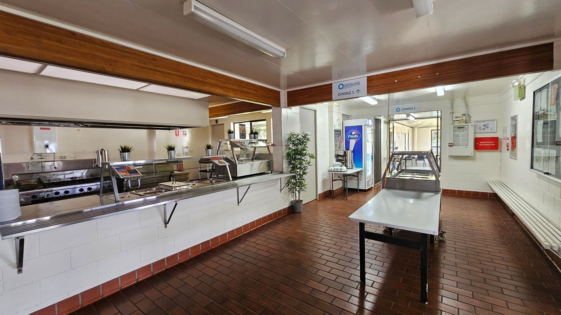 On the left, there is a serving counter with metal fixtures and several trays arranged neatly, alongside potted plants for decoration. A small window reveals more kitchen space behind the counter. To the right, a vending machine and a table with hot drink dispensers are positioned against the wall. In the middle, a stainless steel table stands ready for food preparation. The floor features dark brown tiles arranged in a checkerboard pattern, while the ceiling is flat with fluorescent lights.