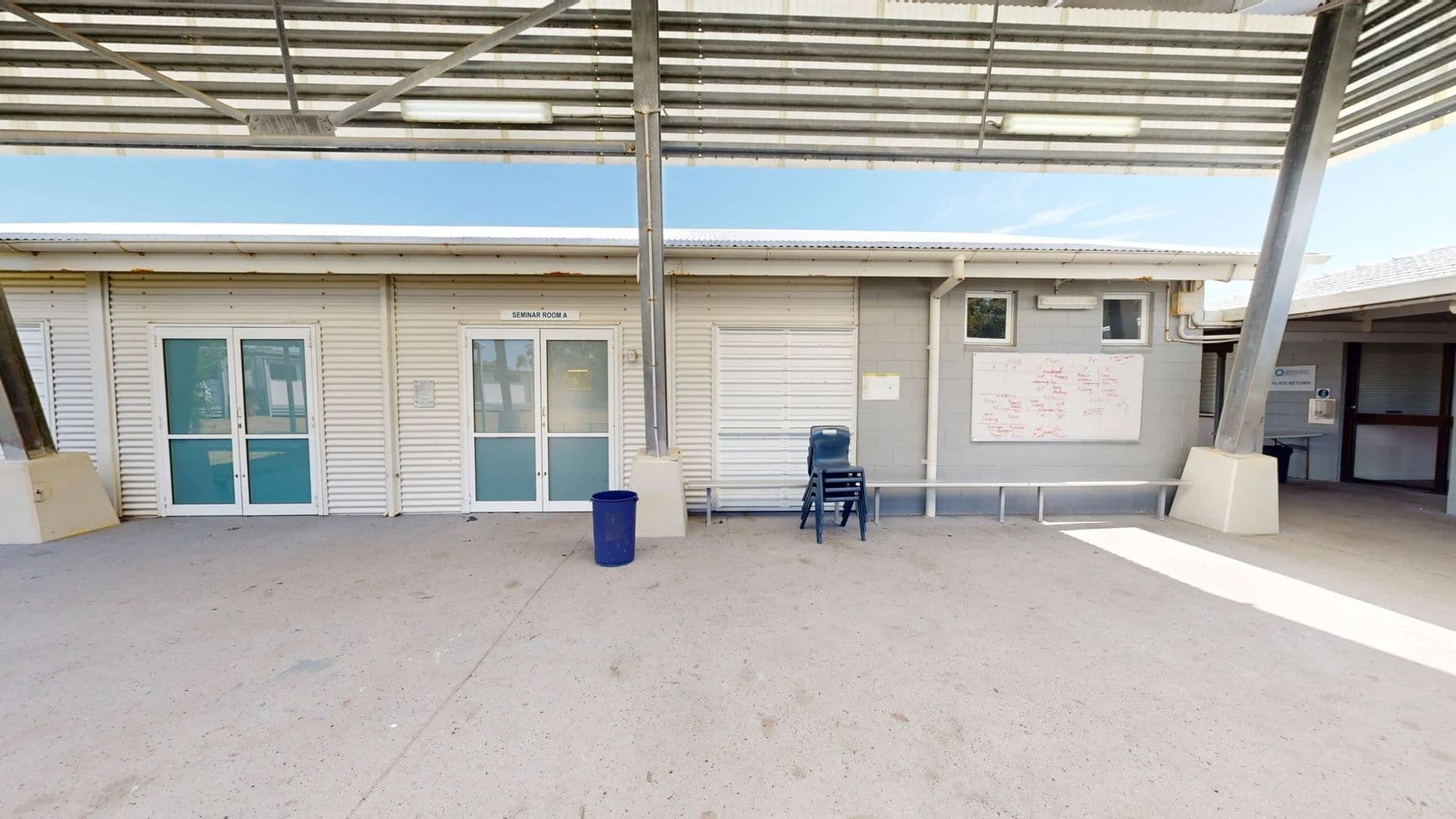 On the left, two glass doors marked "Seminar Room A" lead into a building with a light-coloured, corrugated exterior. Next to it, a wall with a whiteboard covered in notes is visible. To the right, a second entrance leads into a different room, with a small window above it. A blue bin stands on the concrete floor, alongside a stack of chairs. The ground is a smooth, pale concrete surface. Above, a metal roof supported by beams provides shade, with fluorescent lights mounted underneath.