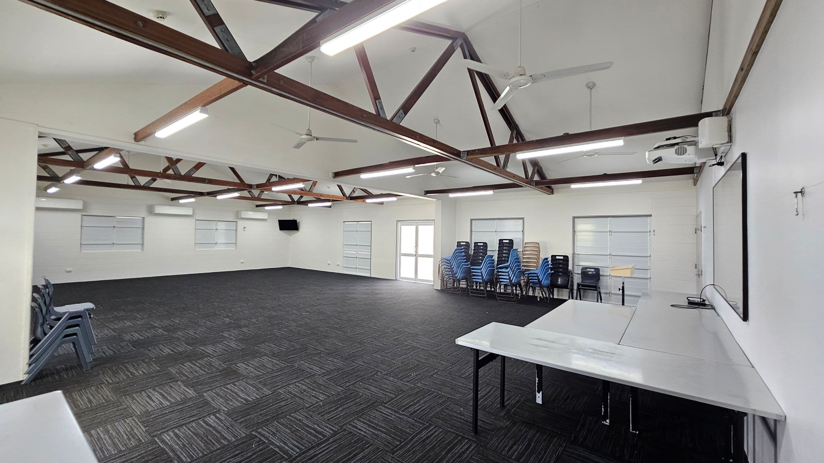 To the left, a stack of grey chairs is arranged against a white wall, beside a doorway. On the right, a row of windows lets in light, with a wall-mounted television just above. A large whiteboard is mounted nearby. In the middle, a rectangular table with a smooth surface is positioned at an angle, facing the room. The floor features dark carpet tiles arranged in a patterned layout. The ceiling has exposed wooden beams, with ceiling fans and fluorescent lights evenly spaced throughout.