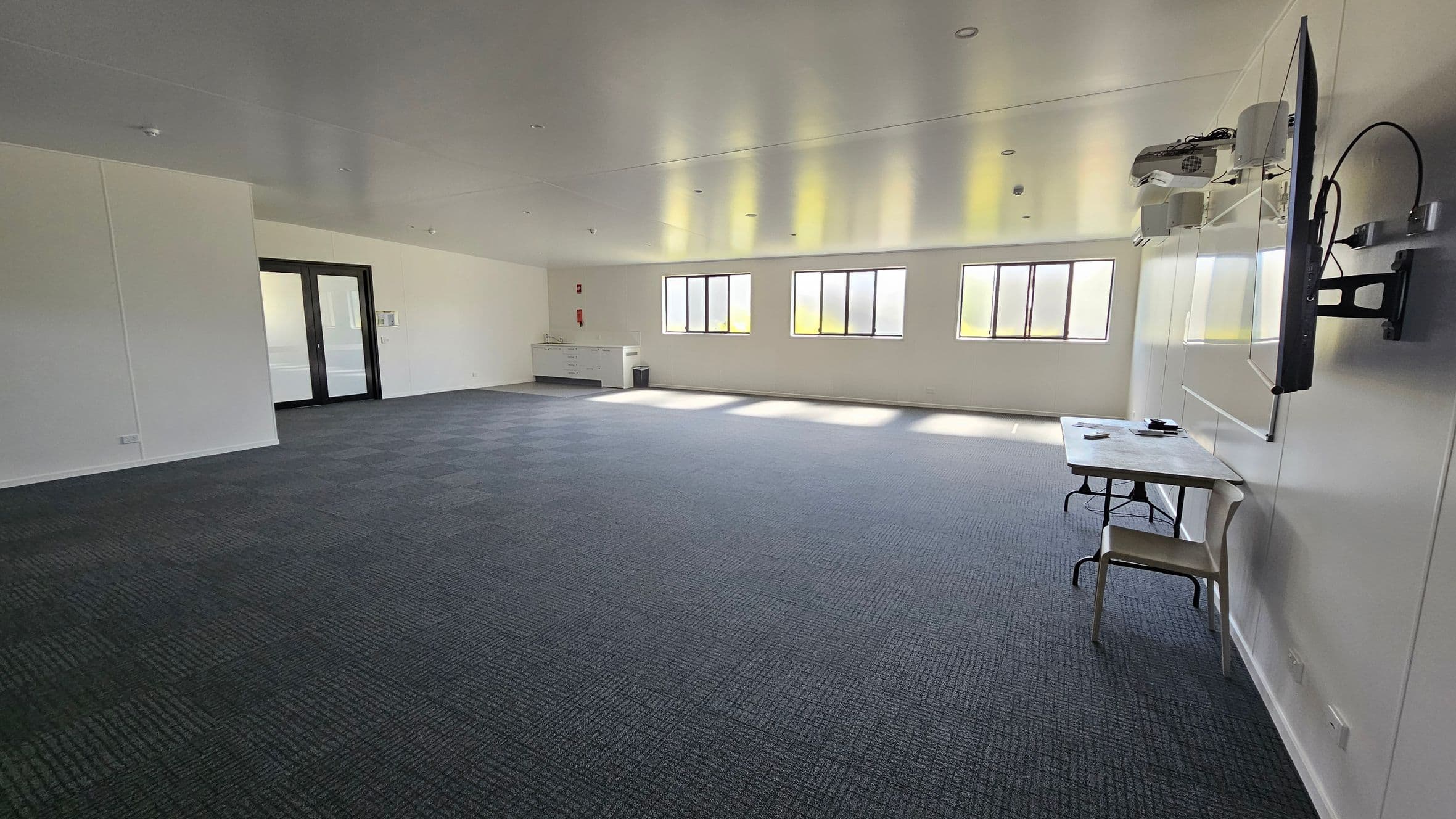 On the left, large double doors lead to another space, with a wall-mounted notice board beside them. A service counter is situated nearby, featuring a sink and storage below. To the right, several tall windows provide natural light, with a flat-screen television mounted on the wall. In the middle, a simple table is set against the wall with a chair beside it. The floor is covered in dark blue carpet with a textured pattern. The ceiling is flat and smooth, fitted with recessed lighting.