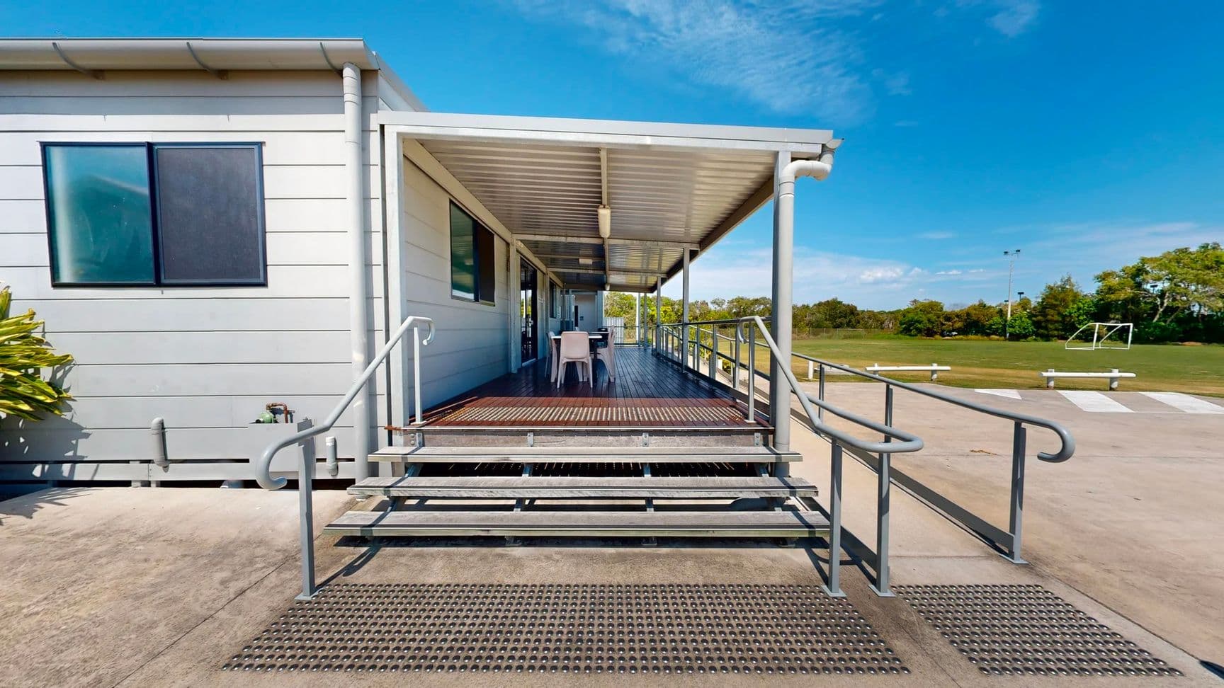On the left, a small window with a dark frame is visible, alongside a light-coloured wall of a building. A cluster of green plants is situated next to the steps. To the right, a set of stainless steel rails leads to a deck area with a table and chairs under a corrugated metal roof. The foreground features a wide, grey concrete pathway with a textured surface, while the background reveals a grassy area with white benches and a goalpost in the distance.