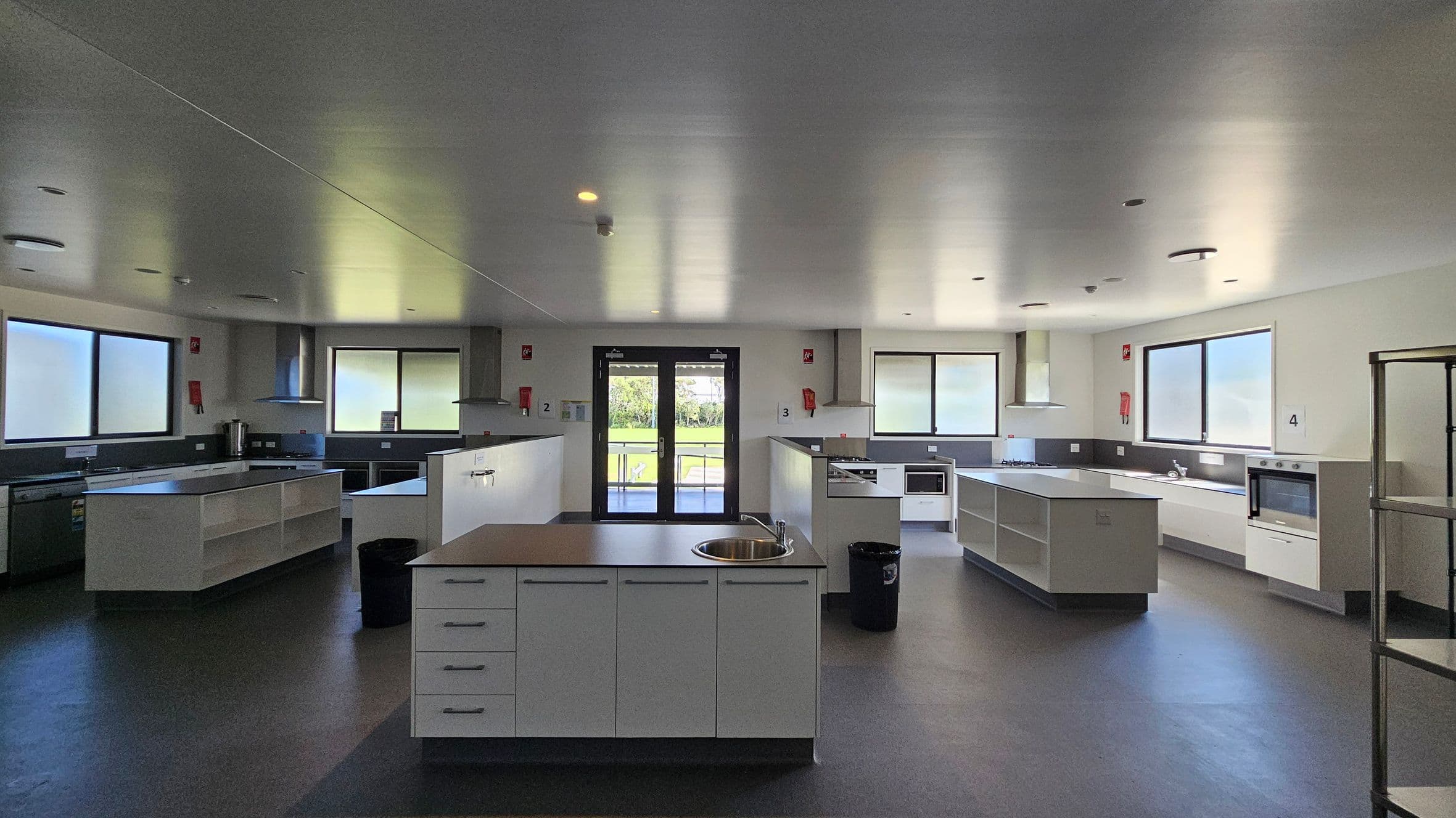 On the left, a stainless steel kitchen unit includes a sink and countertops, with cabinets above for storage. To the right, another cooking station features a hob and an oven, alongside more countertop space. In the middle, an island with a circular sink is surrounded by drawers. The floor is a dark grey, smooth vinyl, while the ceiling is flat and painted white, with recessed lights evenly distributed. Large windows allow light into the room, and a set of double doors leads outside.