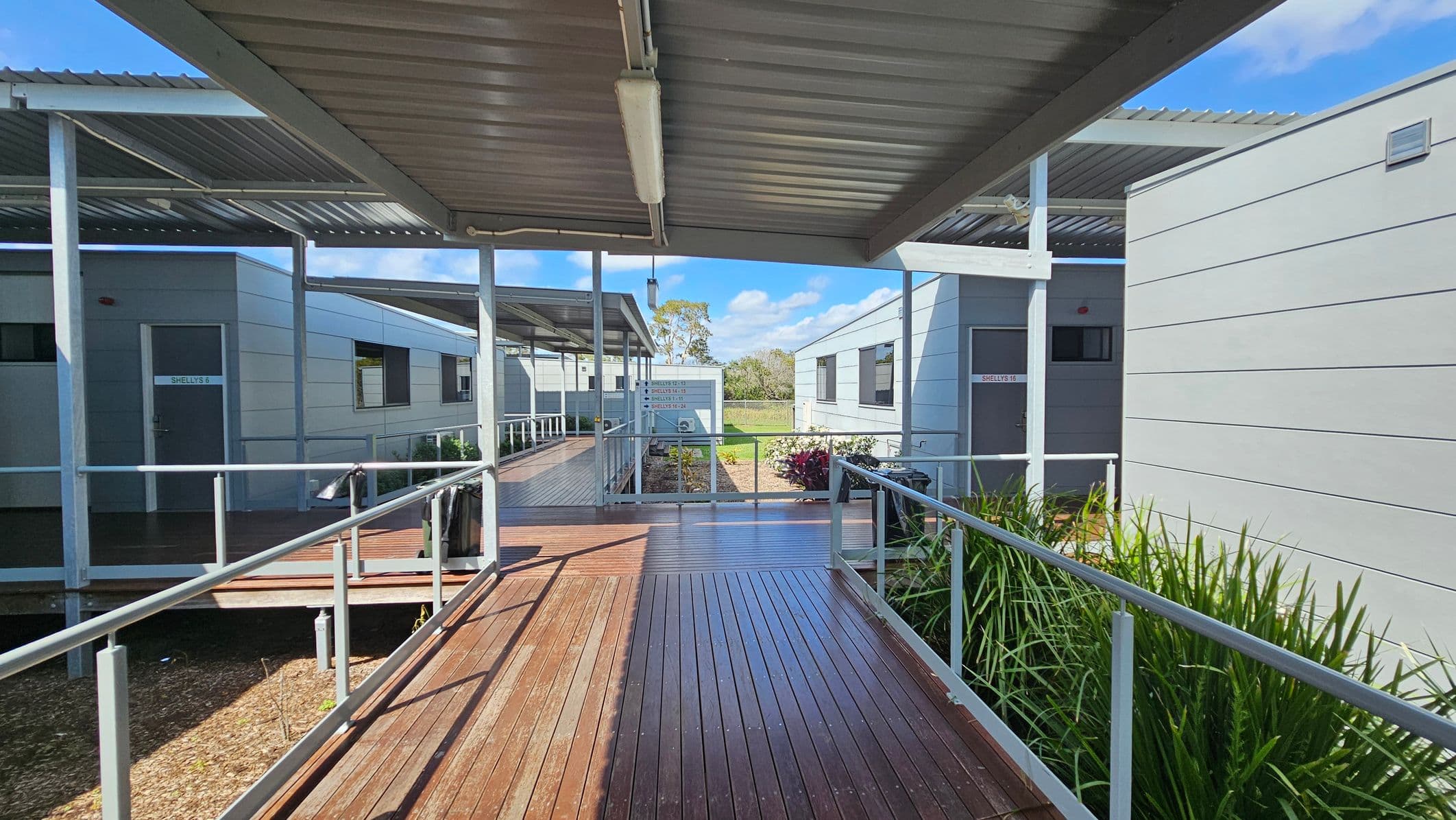 Covered walkways with wooden decking connect several grey modular buildings. On the left, a door labelled "SHELLEY 5" is visible. On the right, another building has a door marked "SHELLEY 16". Metal railings line the walkways, and there are green plants along the edges. The ceiling is made of corrugated metal. In the background, a grassy area and trees are visible under a blue sky.